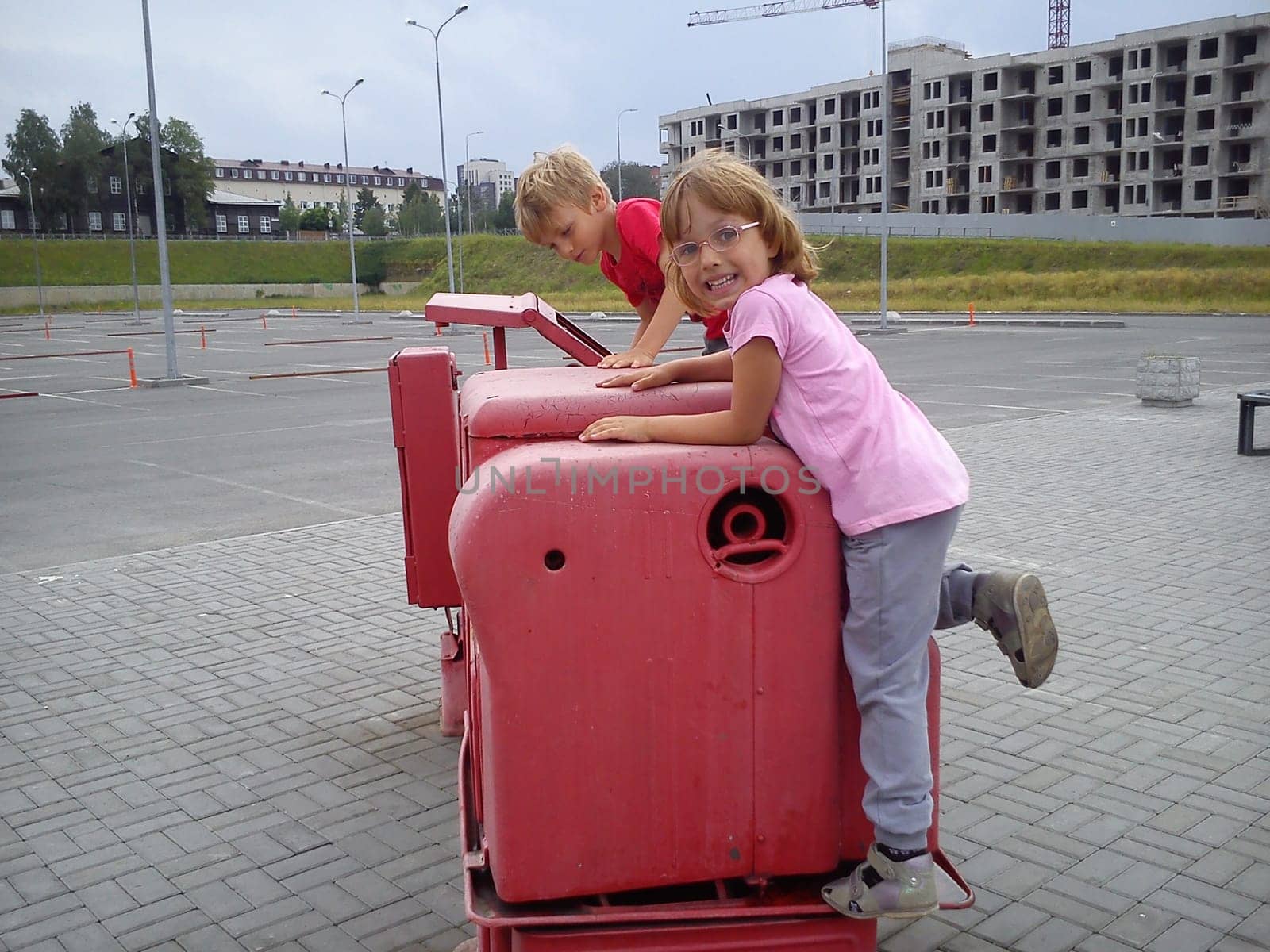 Children climb on the old factory machine, exposed to the street. Factory tractor-machine painted pink paint. Girl laughing boy is serious. Children with blond hair. Construction in the background.
