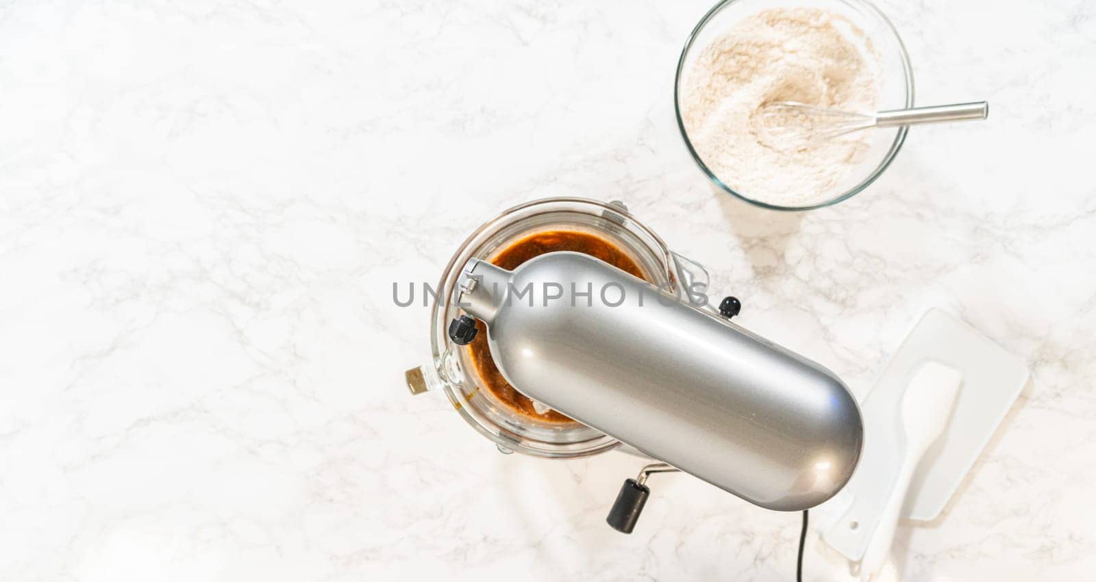 Overhead view of a baking setup for Classic Gingerbread Cookie Dough, with a stand mixer surrounded by essential ingredients. Ingredients include brown sugar, butter, flour mixture, and dark molasses in individual glass bowls, showcasing the classic components needed for this festive dough recipe.