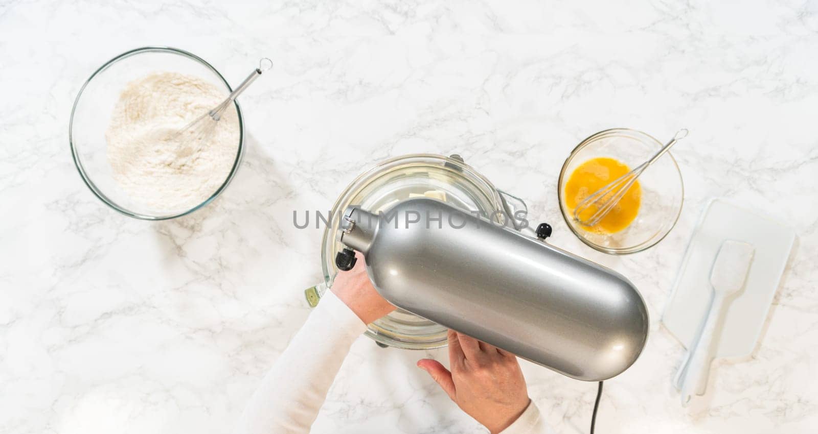 Top view of an electric mixer and bowls containing ingredients for Classic Sugar Cookie Dough. The mixer is placed over a bowl with partially mixed wet ingredients, while nearby bowls hold flour and a beaten egg mixture on a light marble surface.