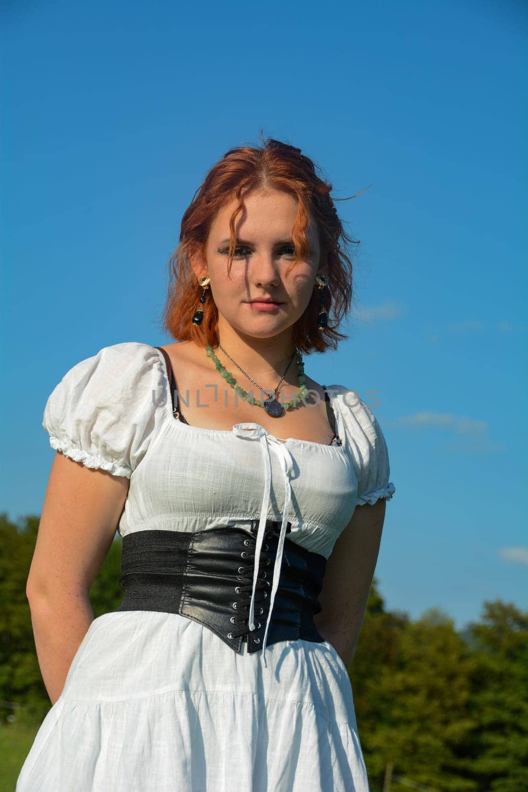 Portrait of a pretty young woman wearing white top, standing outdoors with green trees and blue sky