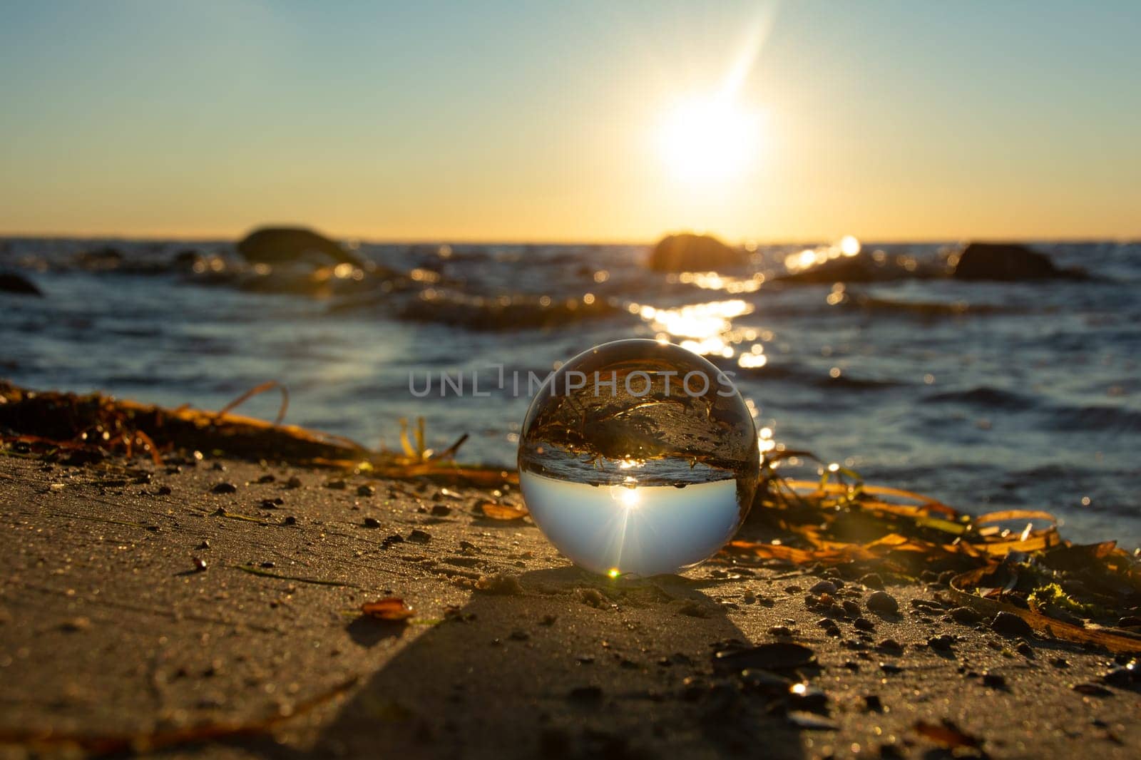 A glass ball lies in the sand in front of the waves, beach with algae and the sea and the setting sun are reflected in the ball