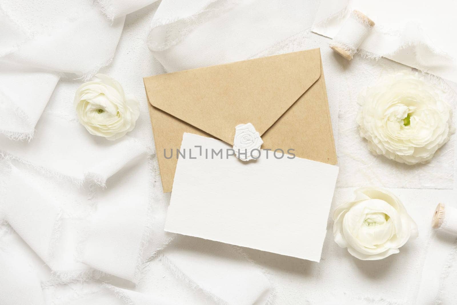 Blank card and envelope near cream roses and white silk ribbons top view, wedding mockup. Romantic scene with vertical card and pastel flowers flat lay. Valentines, Spring or Mothers day concept