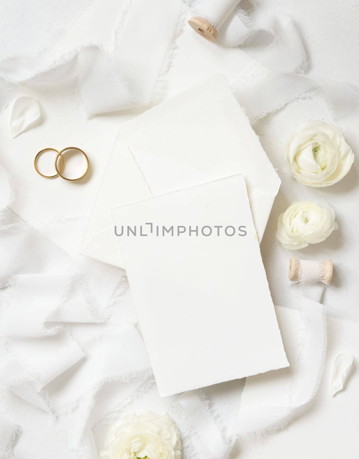 Blank card and envelope near cream roses, white silk ribbons and wedding rings top view, wedding mockup. Romantic scene with vertical card and pastel flowers flat lay. Valentines, Spring or Mothers day concept