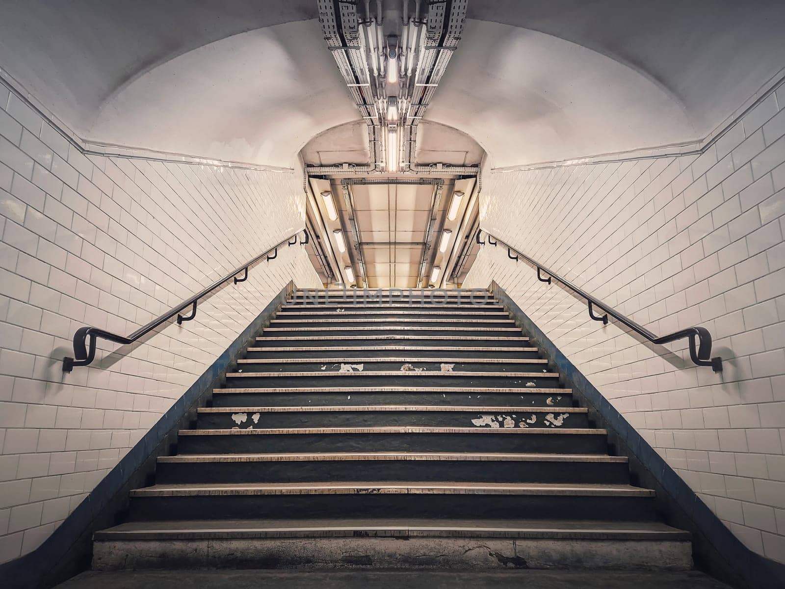 Subway corridor with white tile on walls and glowing lights above a the stairs passage. Symmetrical underground staircase