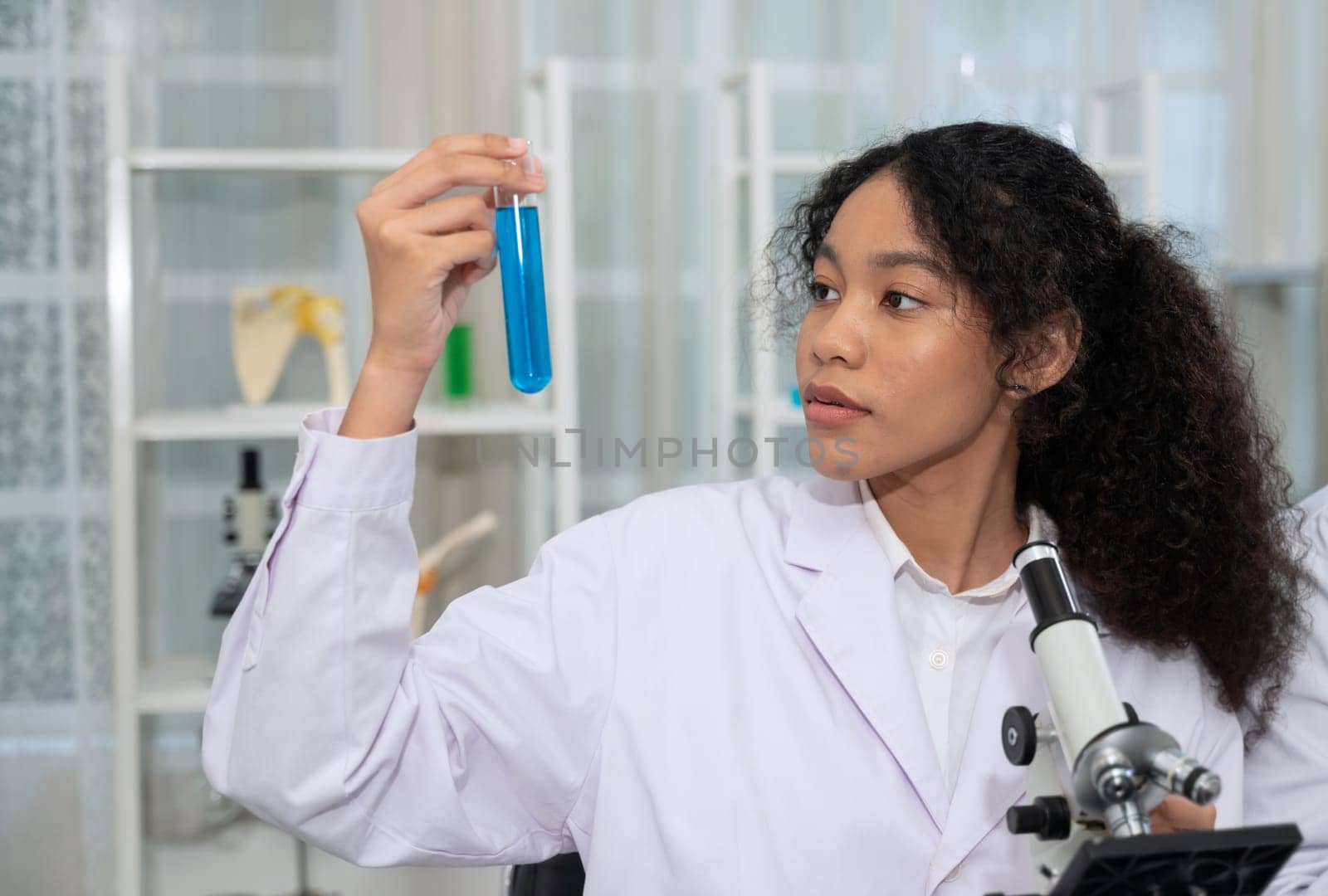 Scientist wearing white lab coat in reserch laboratory. Curly woman holding test tube sample with blue reagent and using microscope