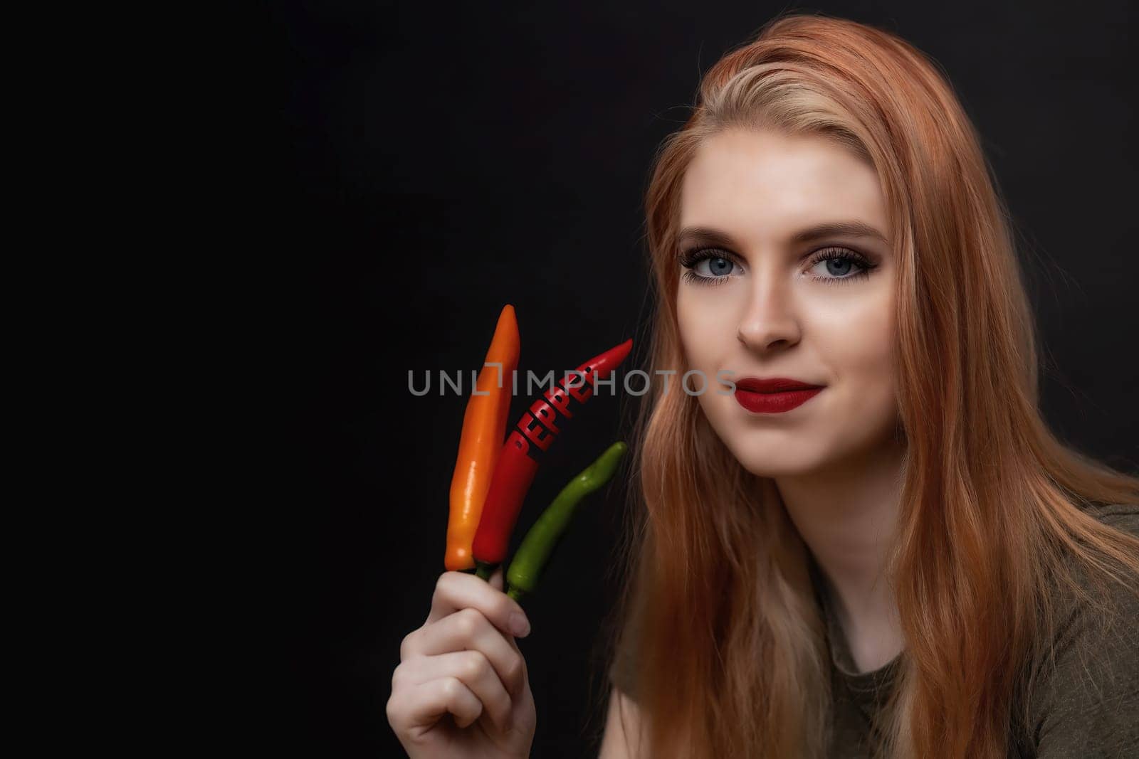 Attractive young woman with red lips is posing with  multicolored chili peppers. One paprika has the inscription paprika carved into it.