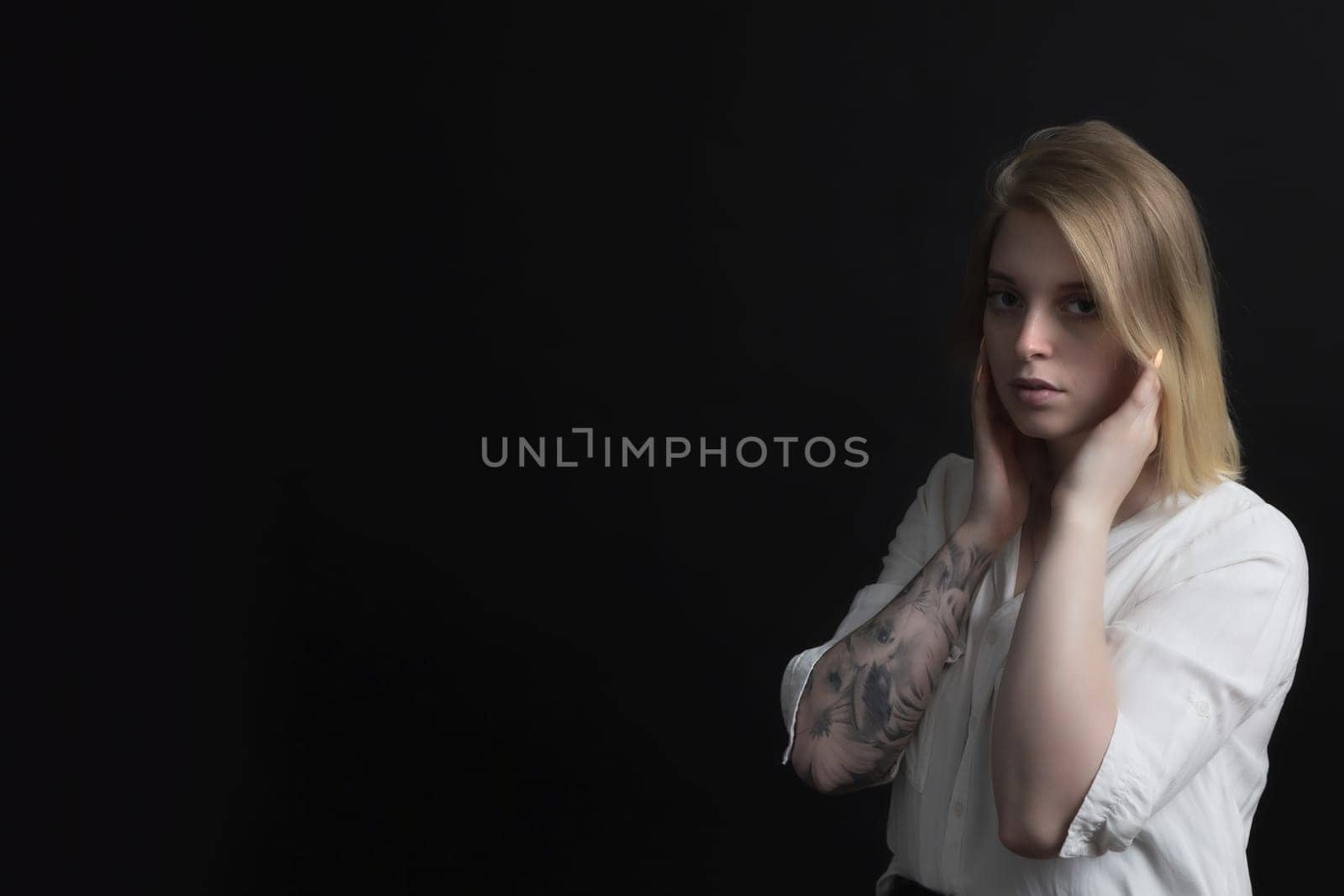 Low key portrait of blonde sad young woman weared in white shirt with tattoo on her hand.