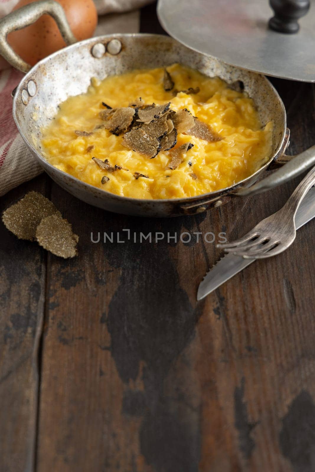 Scrambled eggs with sliced fresh black truffles from Italy served in a a frying pan near fork and knife on wood, close up, copy space. Italian cousine, gourmet breakfast