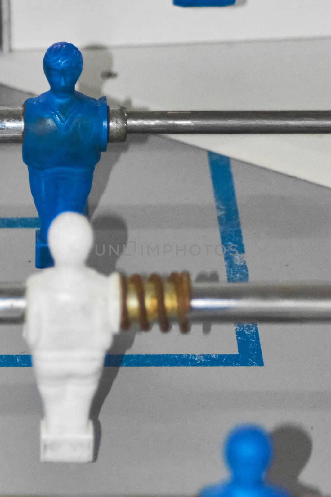 Close-up of foosball player figures, one white and one blue, mounted on metal rods with gold springs. The scene captures the dynamic and fun nature of table soccer games.