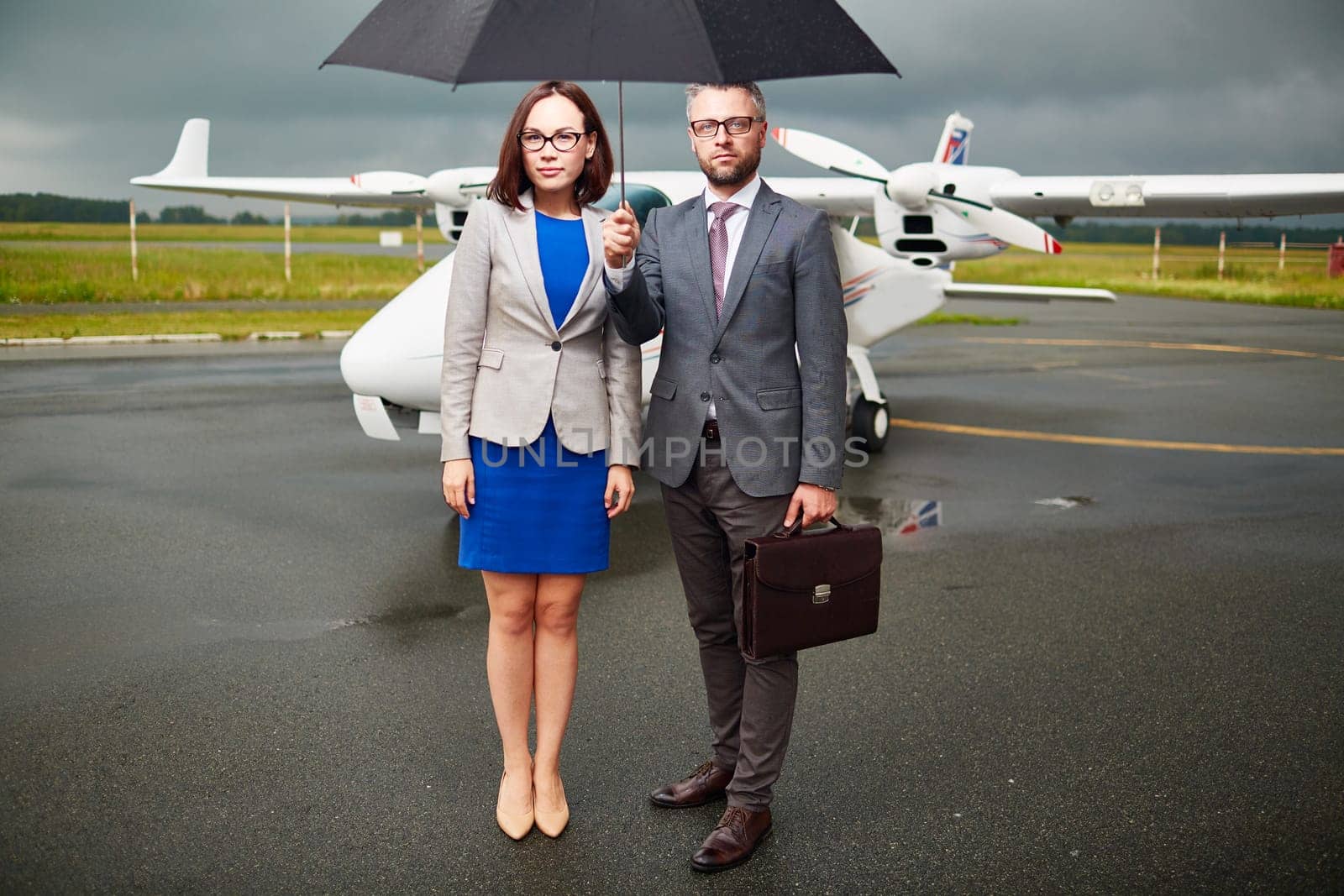 Young businessman holding umbrella over himself and his partner on background of airplane