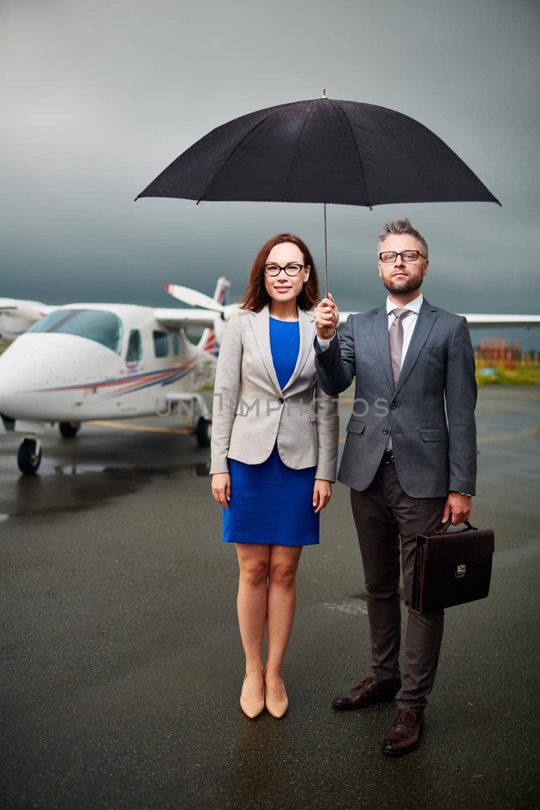 Two confident colleagues under umbrella looking at camera on overcast day