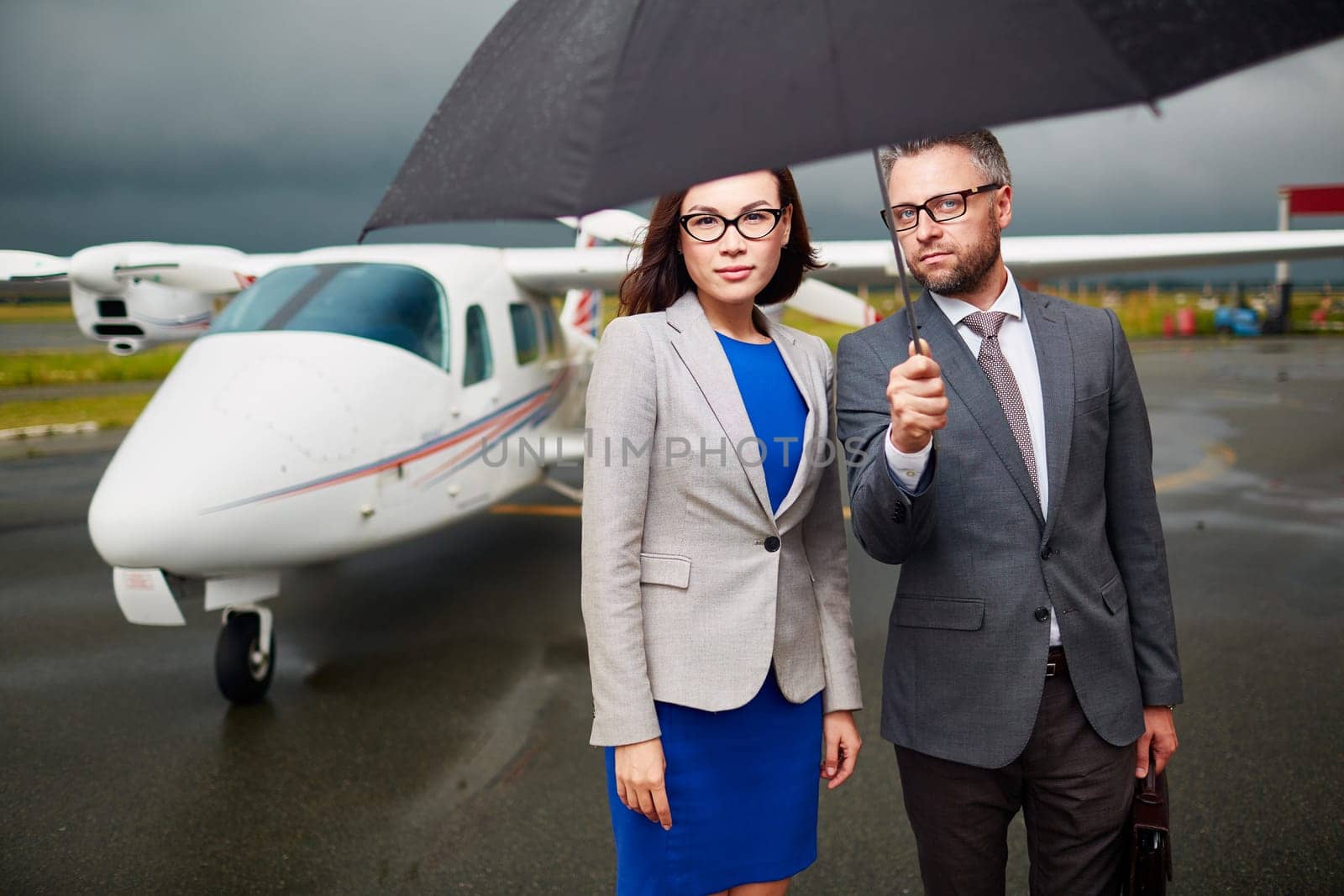 Elegant employees under umbrella looking at camera on rainy day on background of airplane