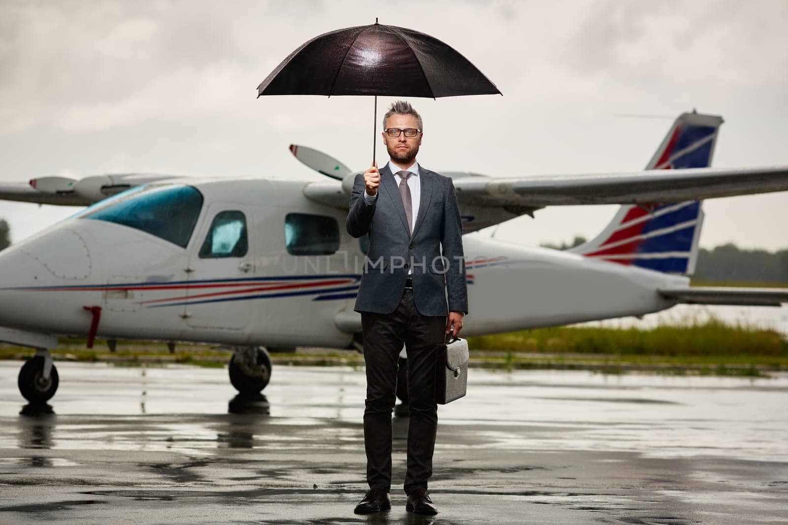 Serious businessman standing on asphalt on overcast day under umbrella