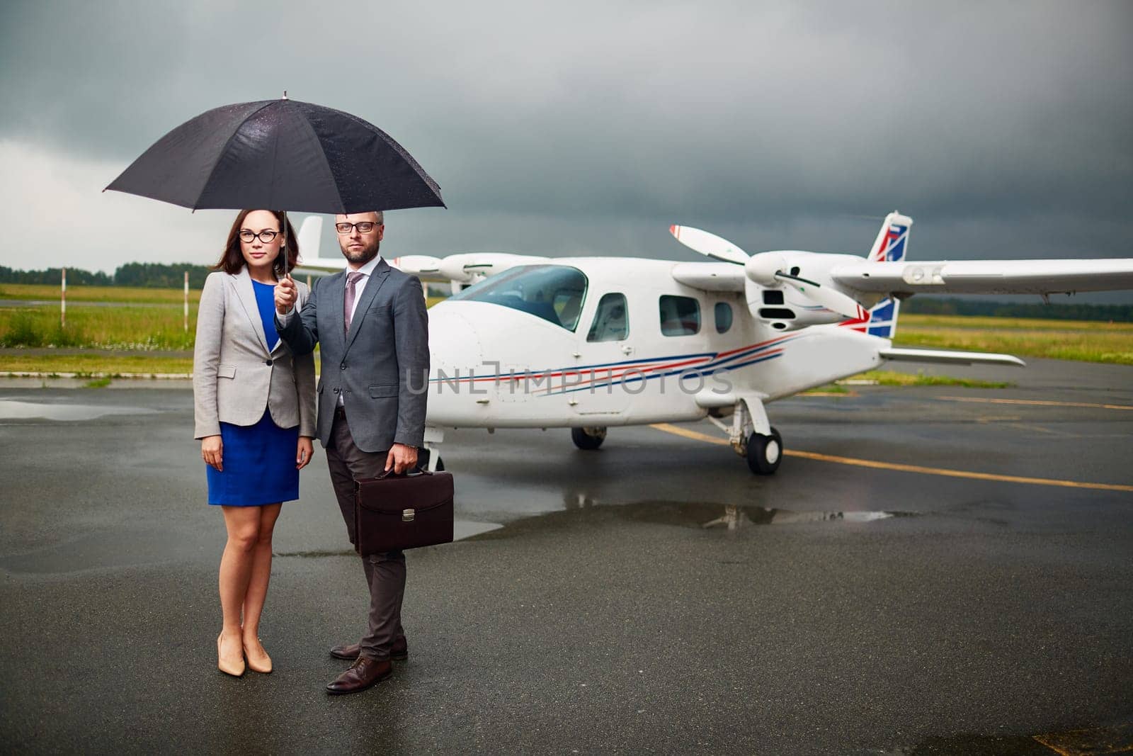 Elegant businessman holding umbrella over himself and his partner with airplane on background