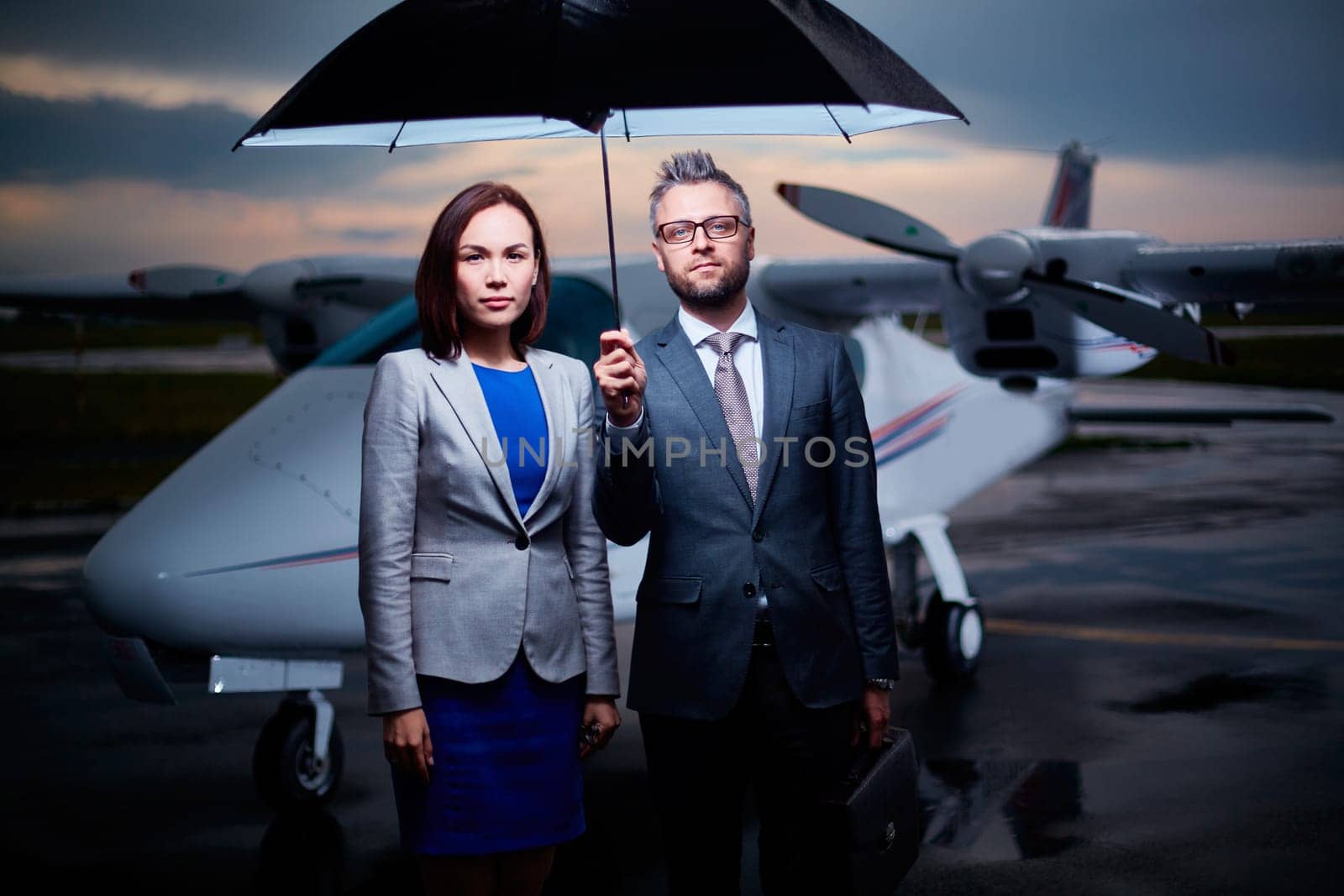 Two confident colleagues under umbrella looking at camera on overcast evening