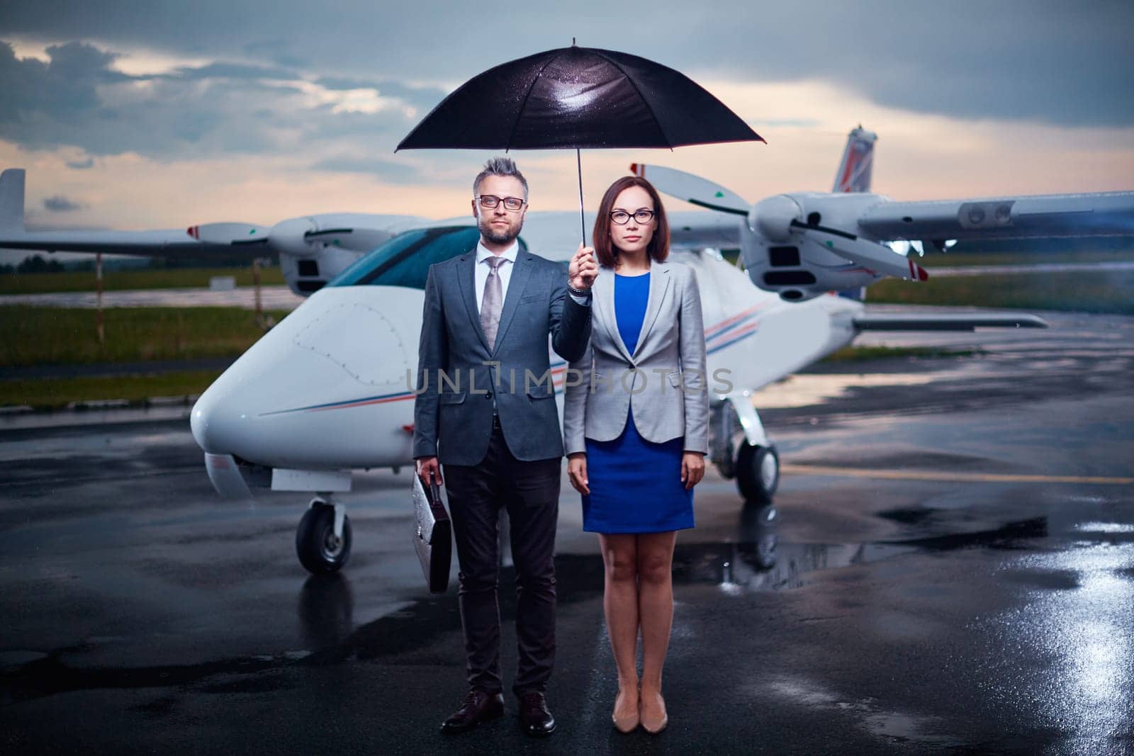 Serious business partners under umbrella looking at camera on rainy evening