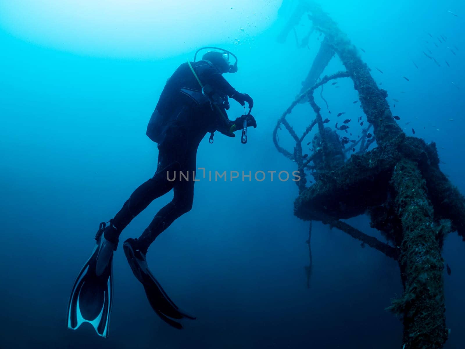 man in a black wet suit is diving a wreck into the ocean, the water is blue and the man is surrounded by fish