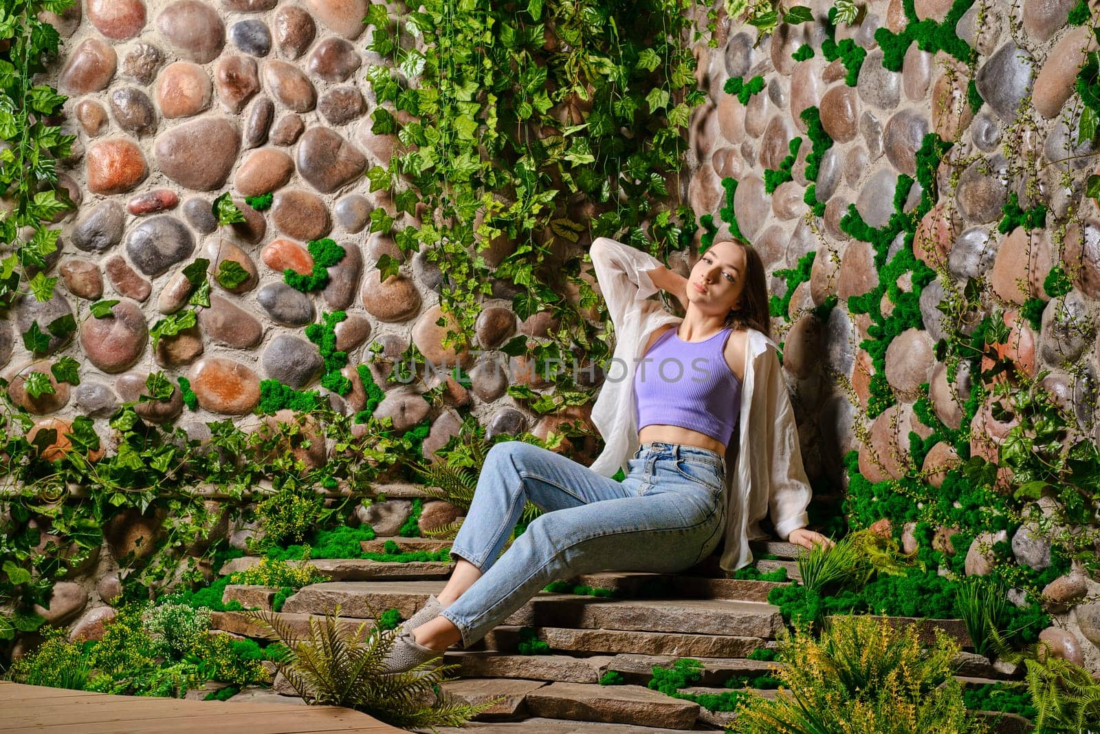 Cute young woman sits on stone plate steps with old overgrown stone wall in the park