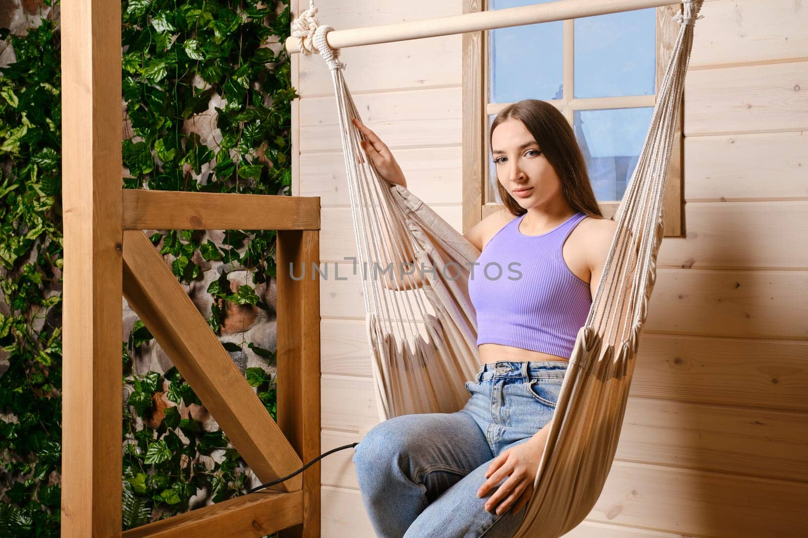 Cheerful woman is sitting in a hammock on the terrace of her country wooden house