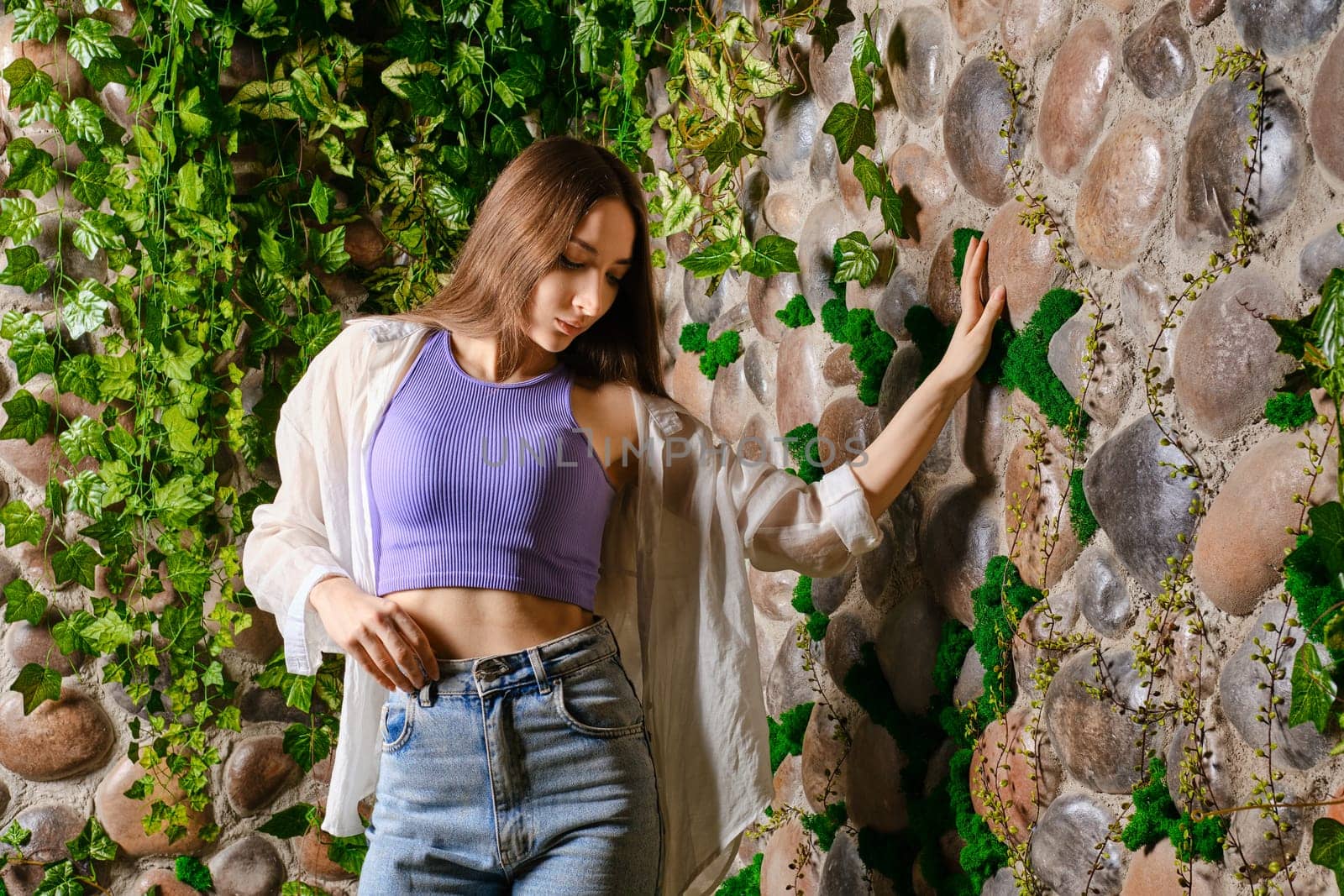 Young dreamy woman touching the stone wall overgrown with plants