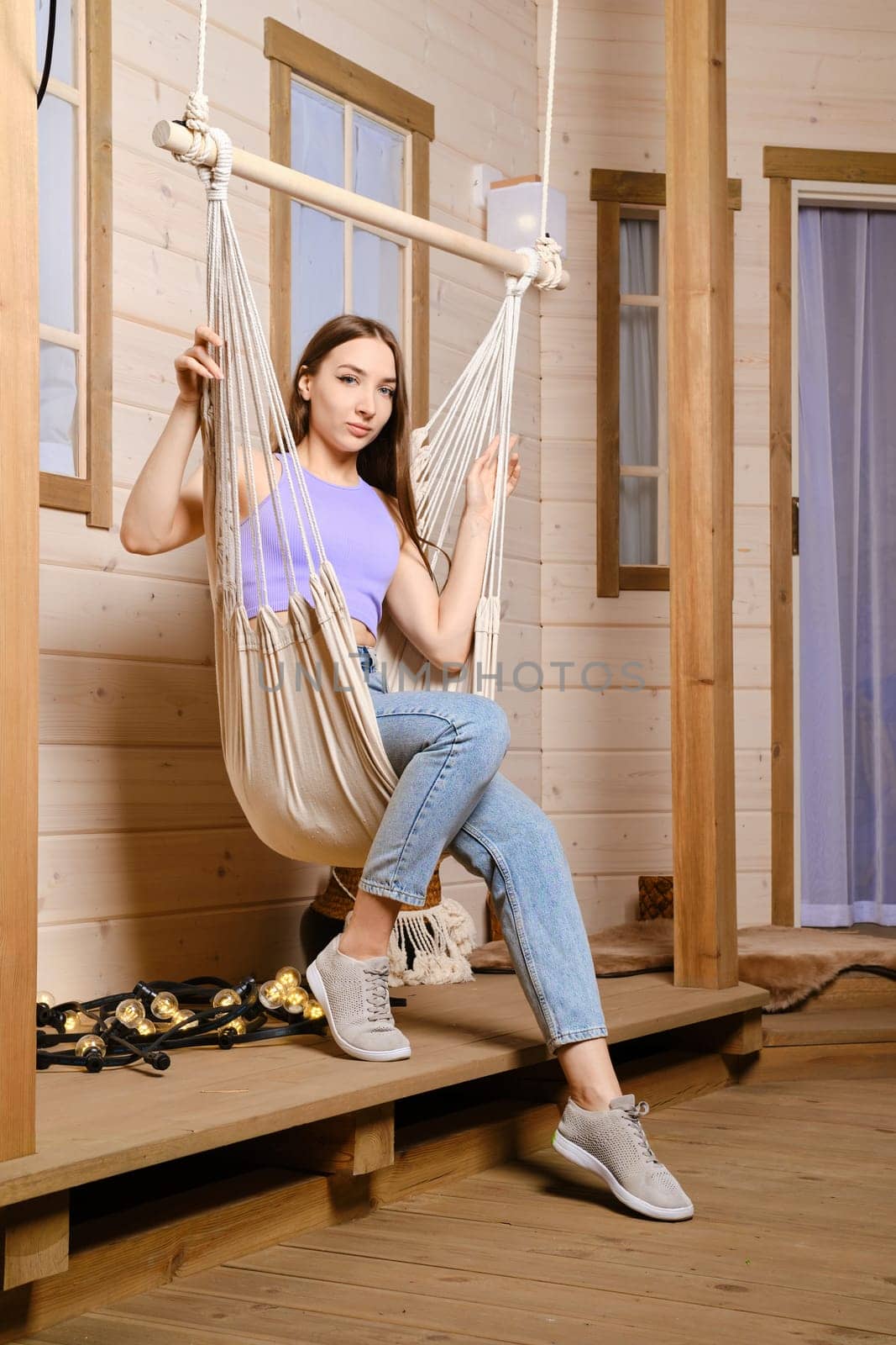 Cheerful woman is sitting in a hammock on the terrace of her country wooden house
