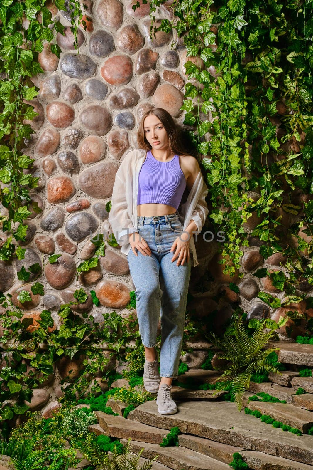 Cute woman leaning on old stone wall overgrown with wild plants