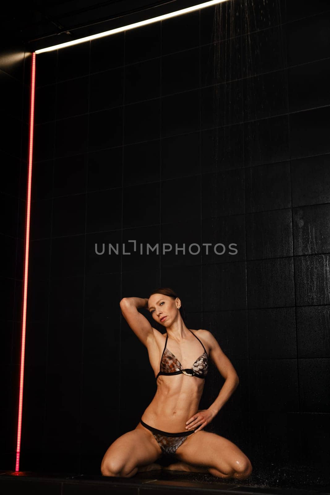 Slender woman is kneeling on the floor in a public shower under running water on black background