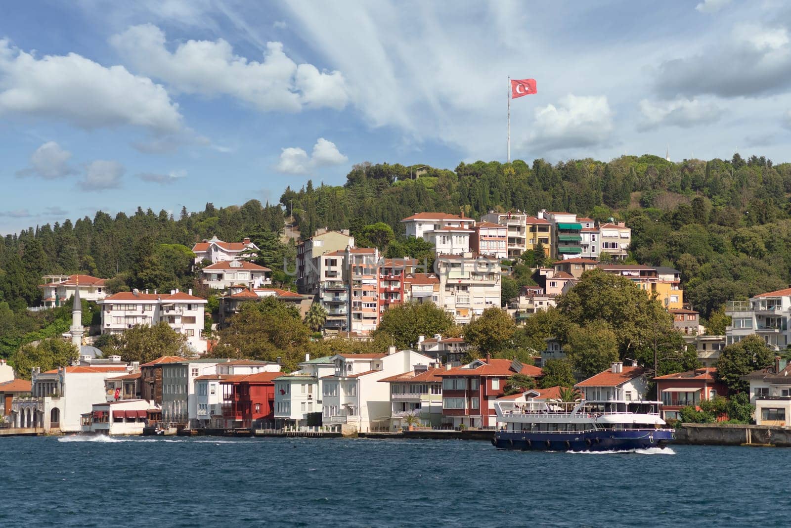 View from Bosphorus strait of the green mountains of the Asian side, with traditional houses, dense trees and a huge Turkish flag in the far end, in a summer day, Istanbul, Turkey