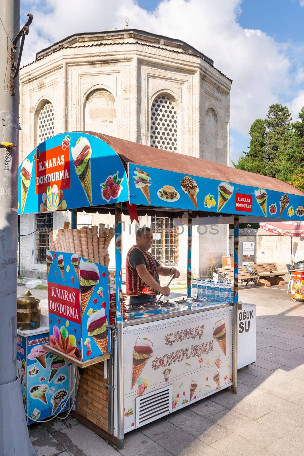 Istanbul, Turkey - September 1, 2022: Middle aged man selling ice cream, or dondurma on traditional Turkish fast food cart with historical tomb in the background at Eyupsultan district
