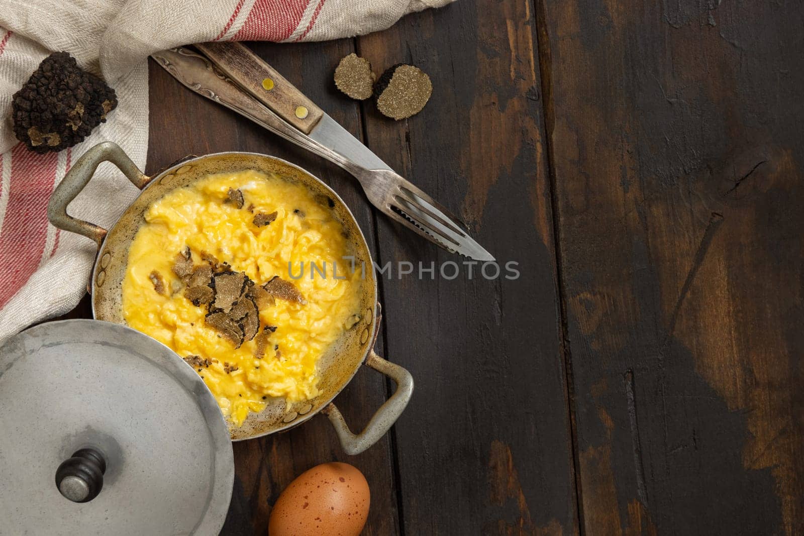 Scrambled eggs with sliced fresh black truffles from Italy served in a a frying pan near fork and knife on wood, top view, copy space. Italian cousine, gourmet breakfast