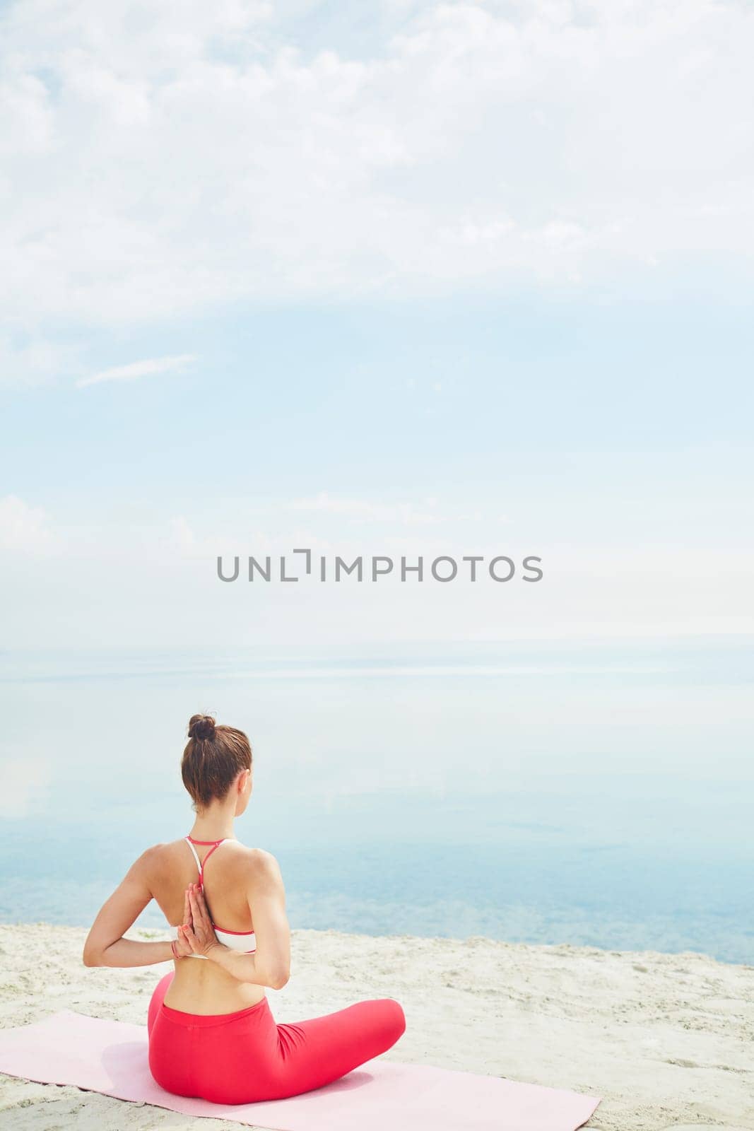 Young woman with crossed legs and arms behind back sitting on sand by water