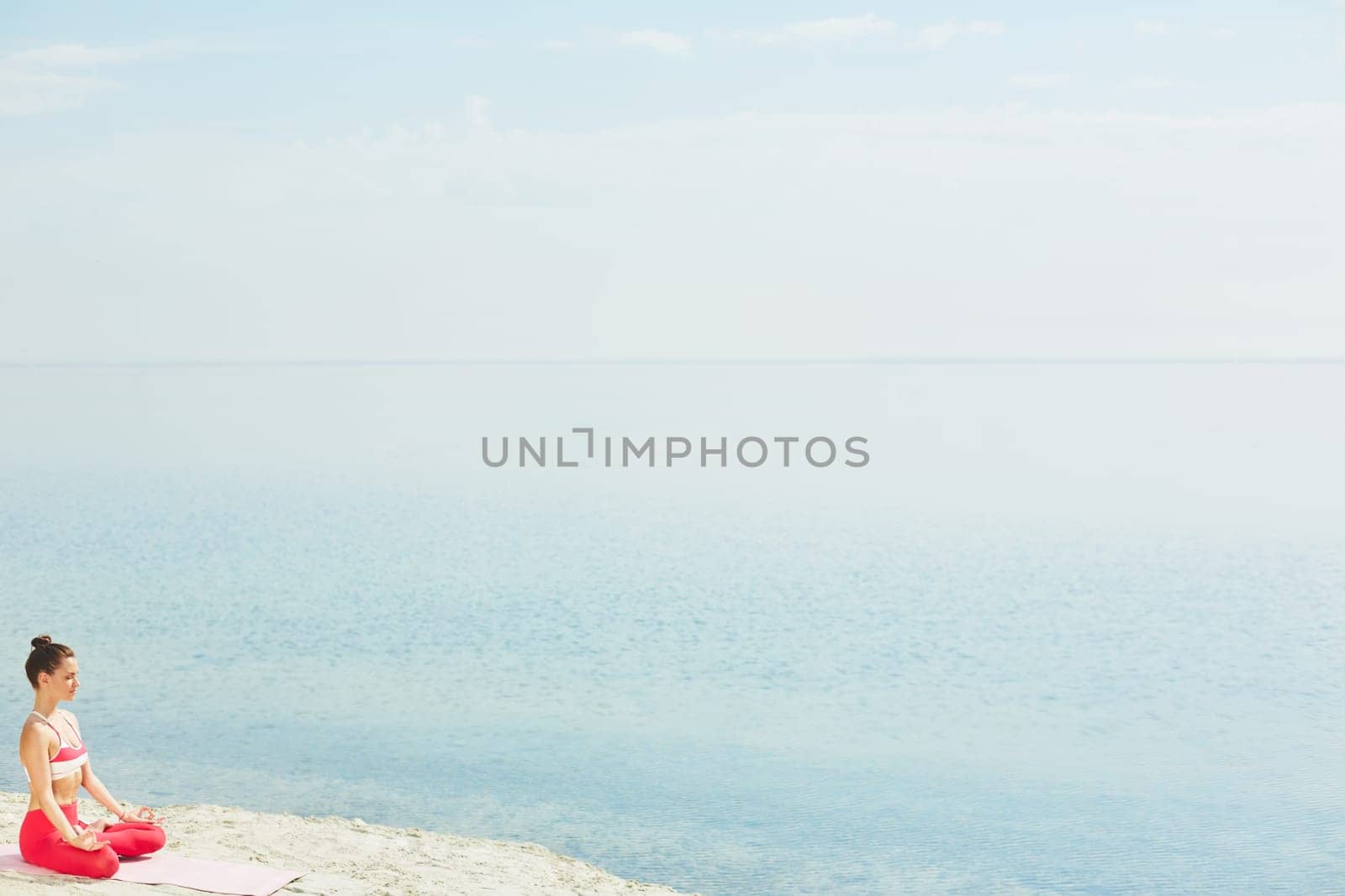 Sporty young woman sitting in pose of lotus on sand by water