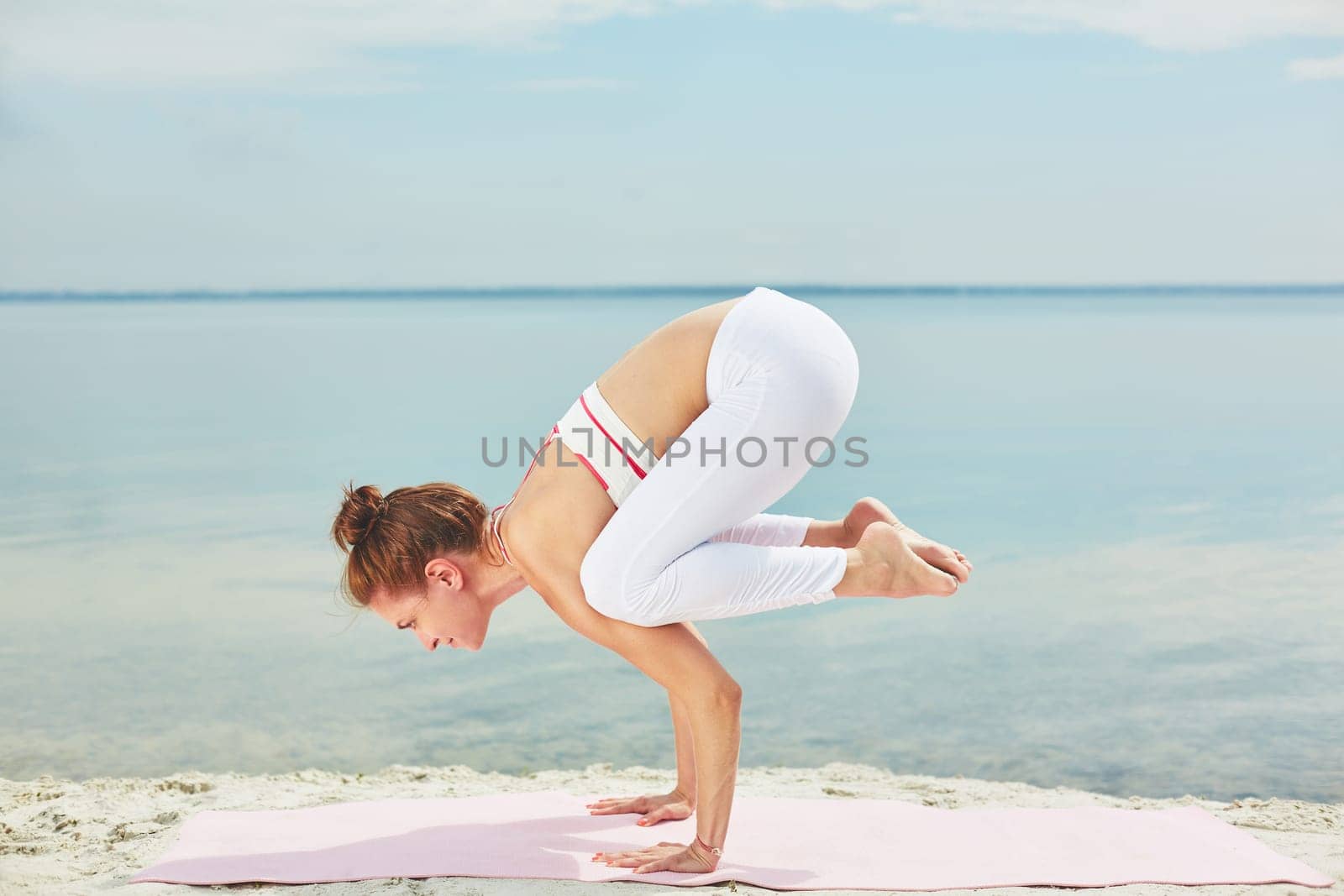 Pretty young woman keeping balance while doing yoga exercise on sand by water
