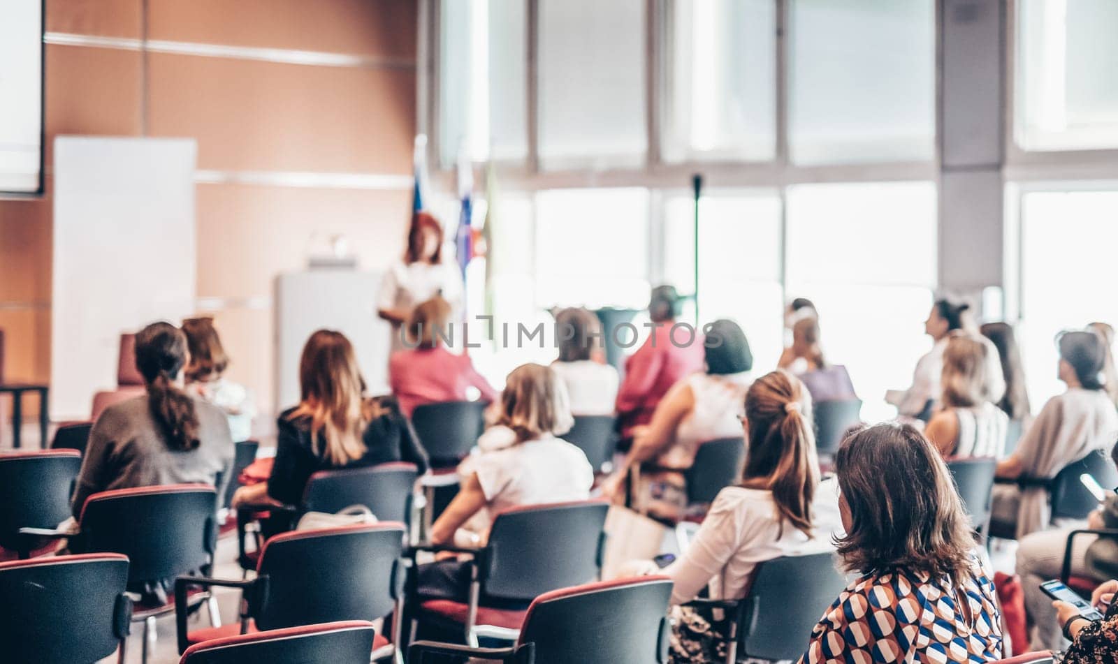 Business and entrepreneurship symposium. Female speaker giving a talk at business meeting. Audience in conference hall. Rear view of unrecognized participant in audience.