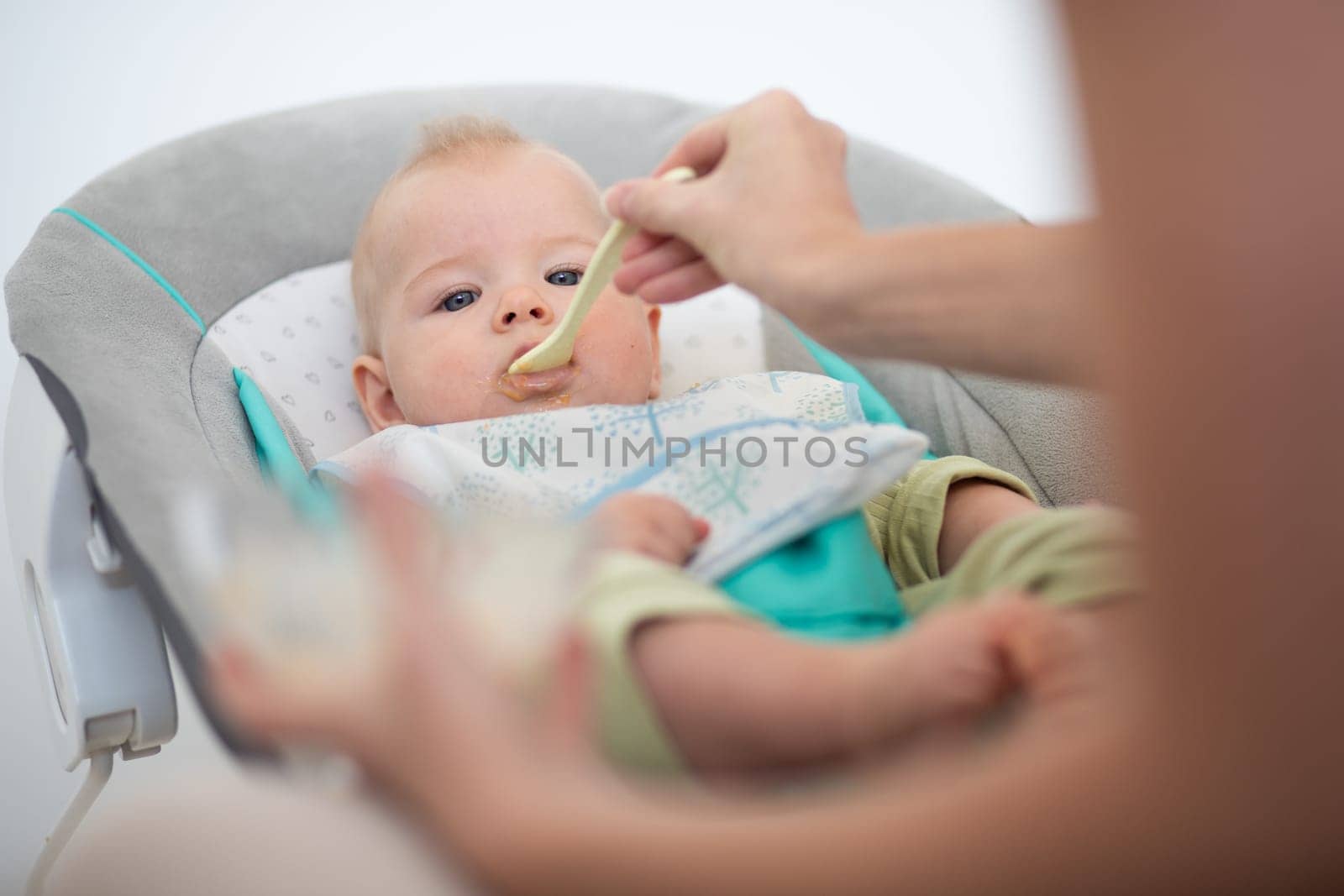 Mother spoon feeding her baby boy child in baby chair with fruit puree. Baby solid food introduction concept