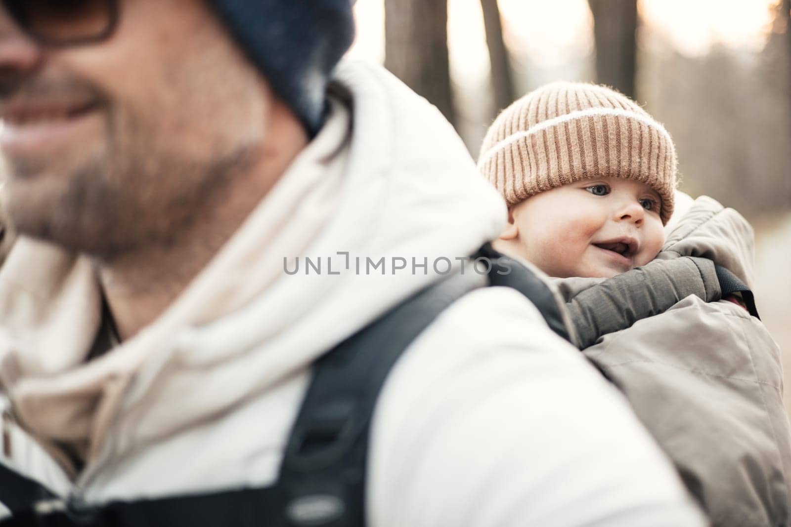 Sporty father carrying his infant son wearing winter jumpsuit and cap in backpack carrier hiking in autumn forest