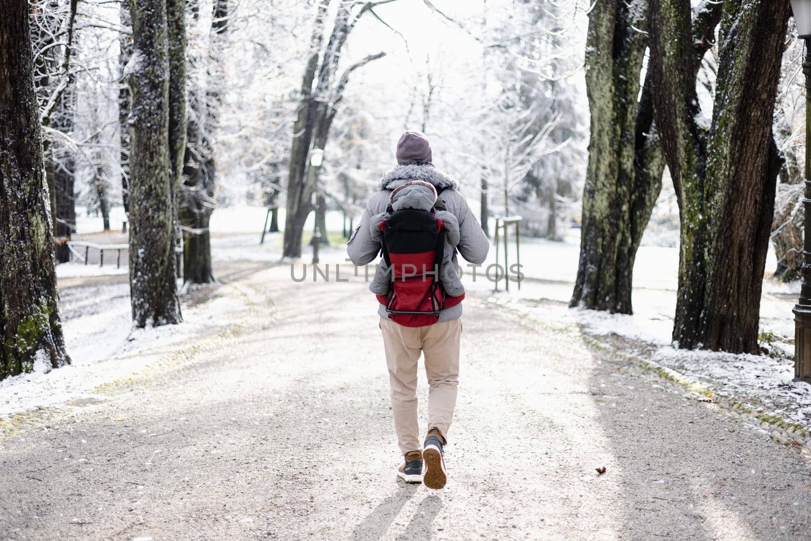 Rear view of sporty father carrying his infant son wearing winter jumpsuit and cap in backpack carrier walking in city park in winter.