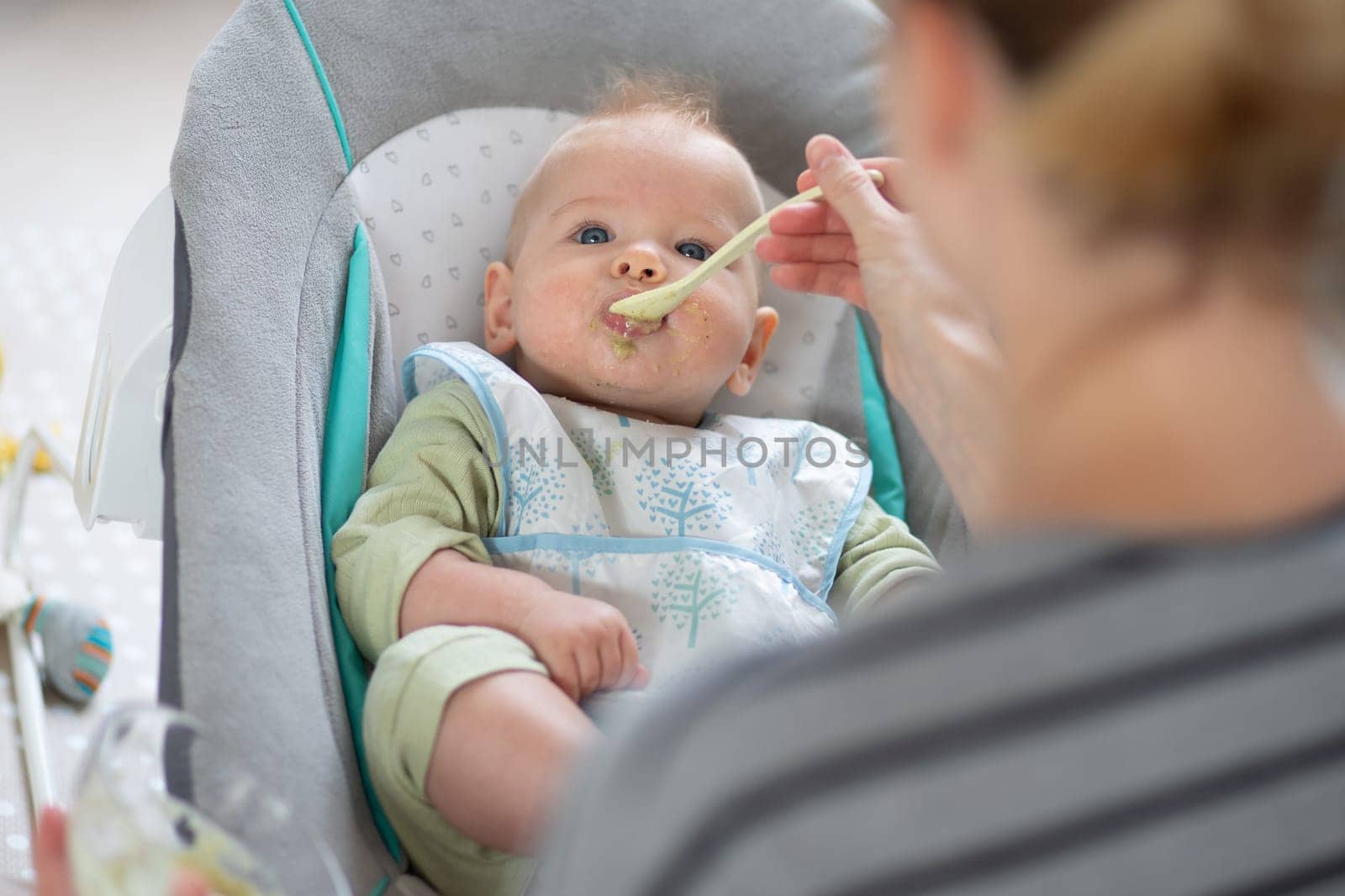 Mother spoon feeding her baby boy child in baby chair with fruit puree. Baby solid food introduction concept