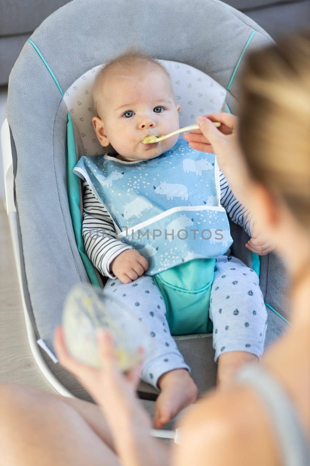 Mother spoon feeding her baby boy child in baby chair with fruit puree. Baby solid food introduction concept