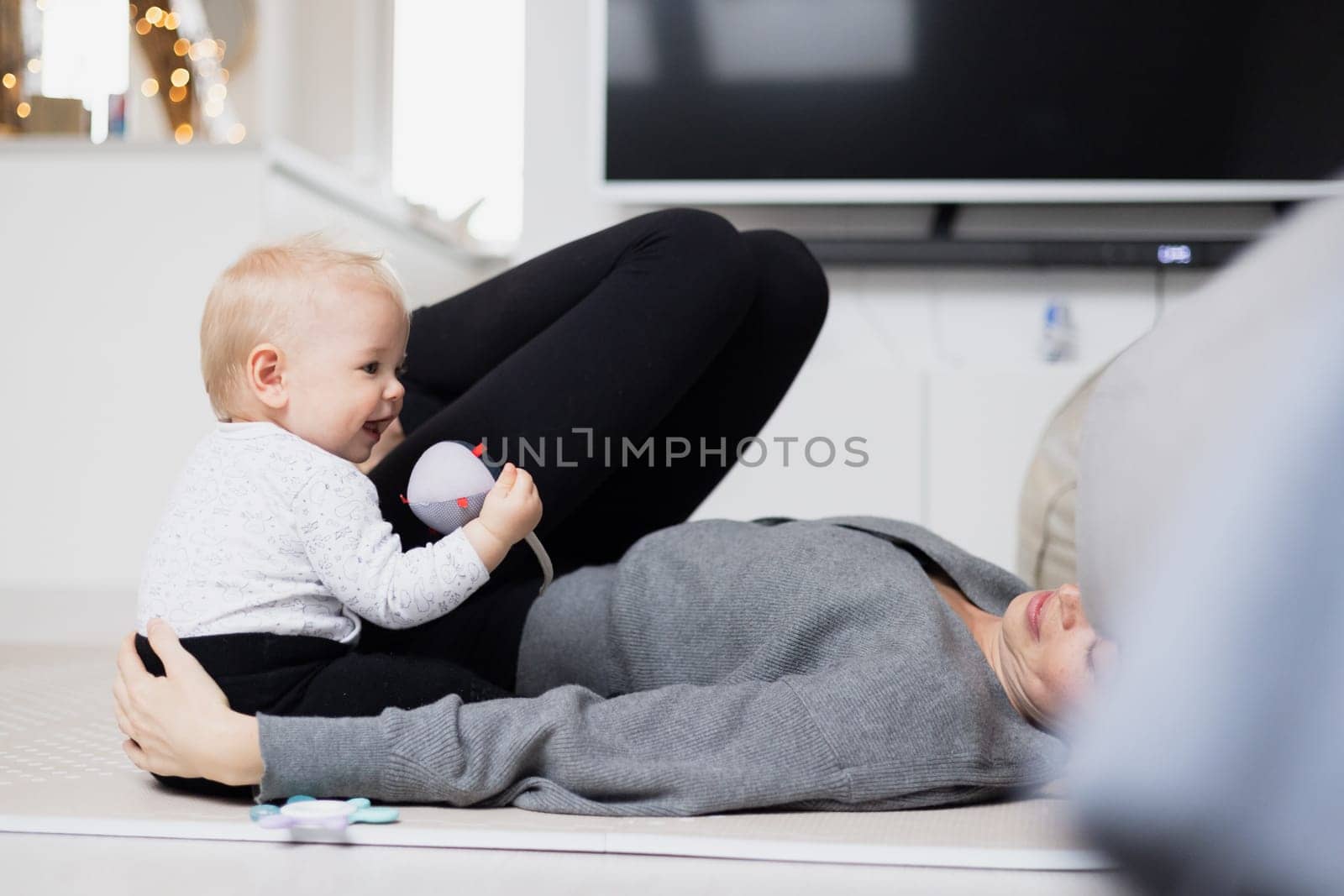 Happy family moments. Mother lying comfortably on children's mat playing with her baby boy watching and suppervising his first steps. Positive human emotions, feelings, joy