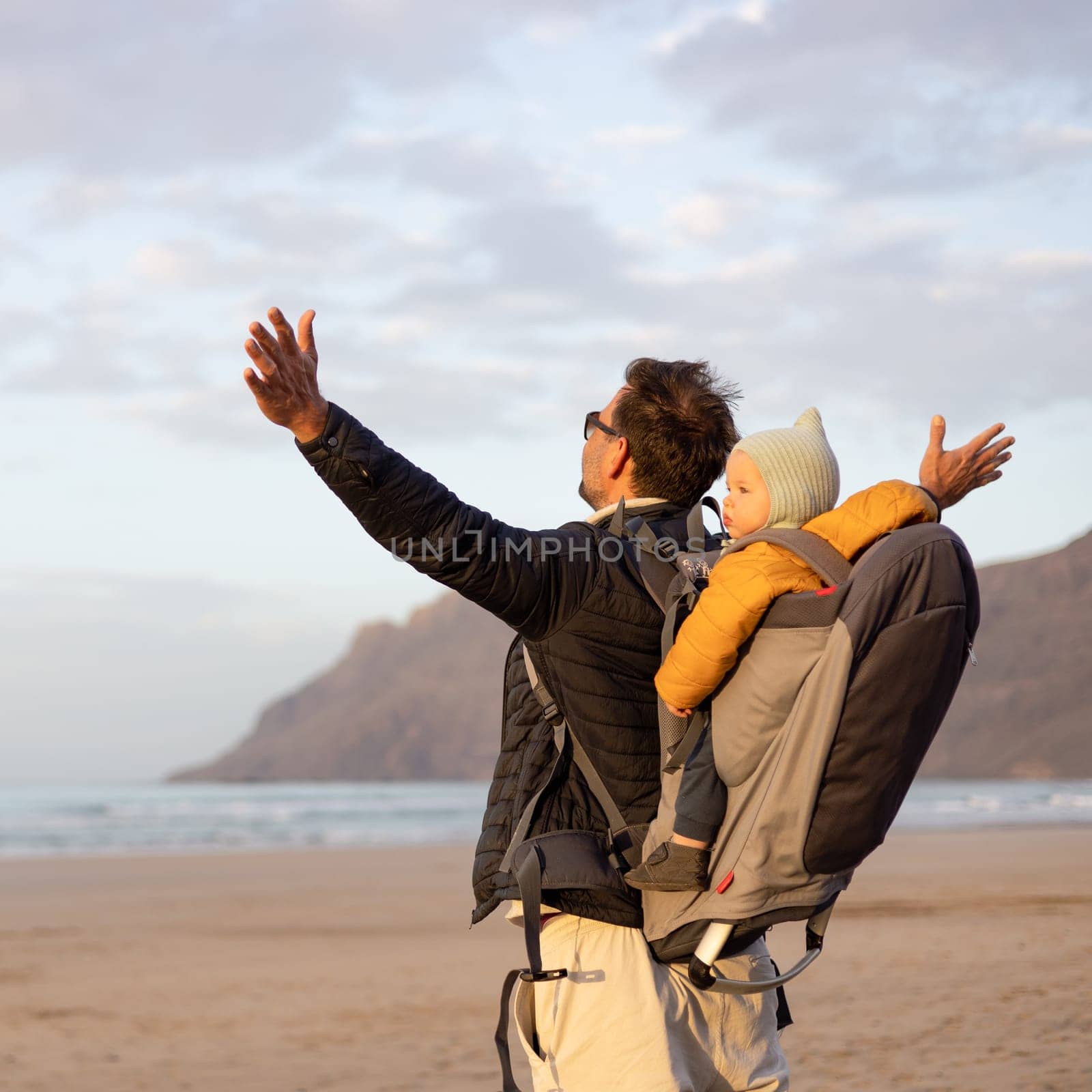 Father rising hands to the sky while enjoying pure nature carrying his infant baby boy son in backpack on windy sandy beach of Famara, Lanzarote island, Spain at sunset. Family travel concept