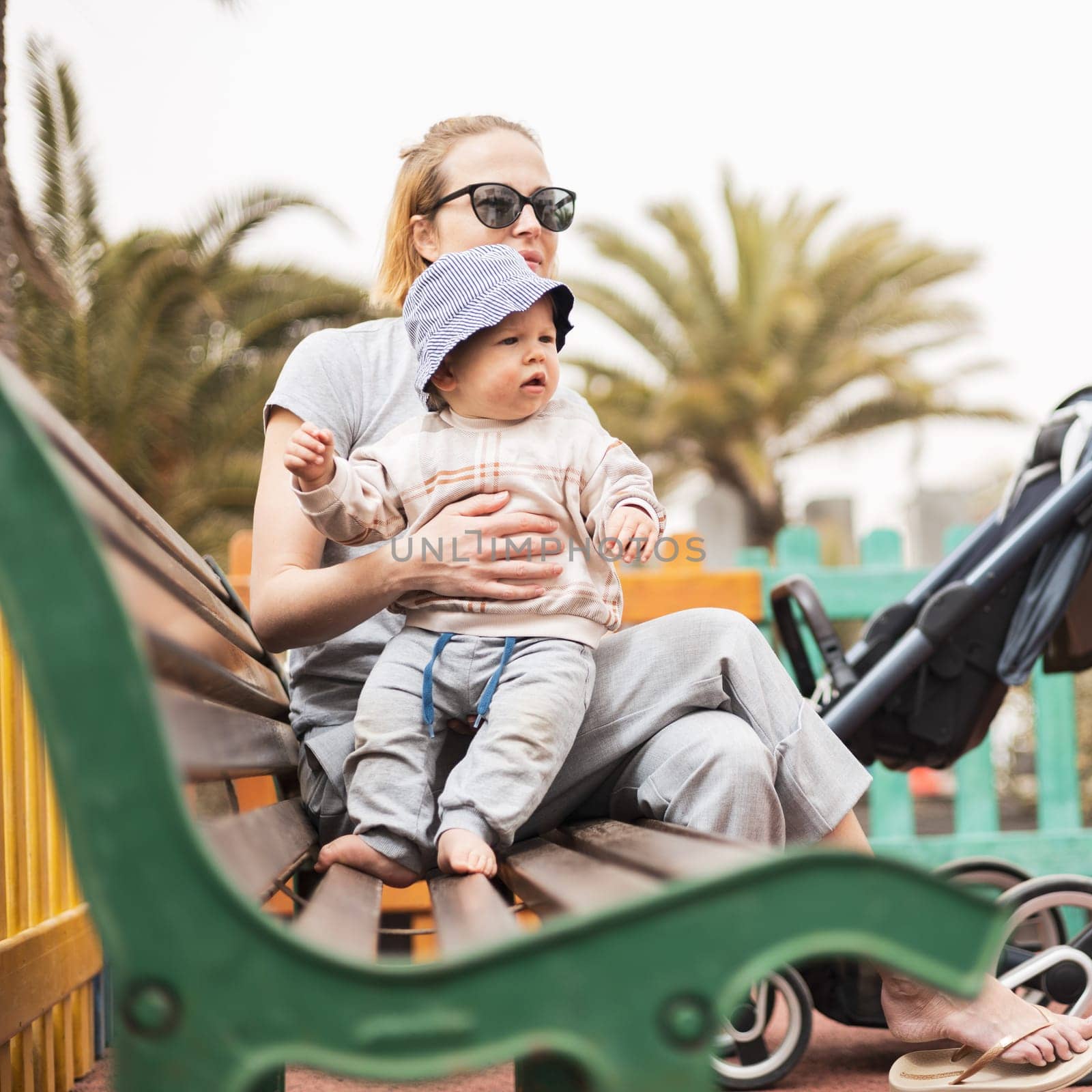 Young mother with her cute infant baby boy child on bench on urban children's playground on warm summer day