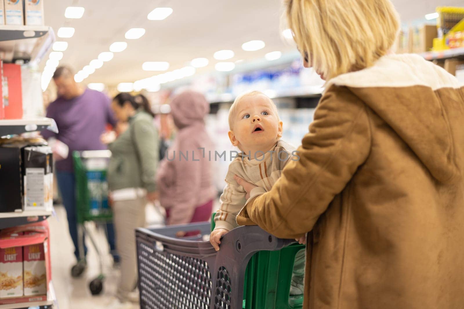 Casualy dressed mother choosing products in department of supermarket grocery store with her infant baby boy child in shopping cart