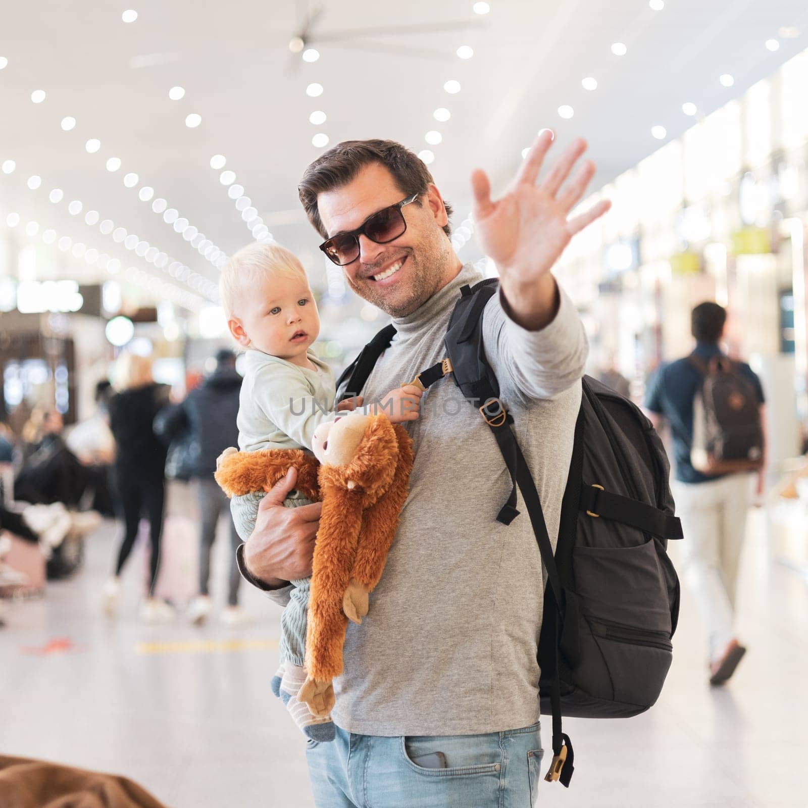 Father traveling with child, holding his infant baby boy at airport terminal waiting to board a plane waving goodby. Travel with kids concept