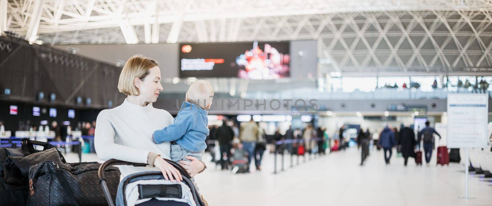 Motherat travelling with his infant baby boy child, walking, pushing baby stroller and luggage cart at airport terminal station. Travel with child concept