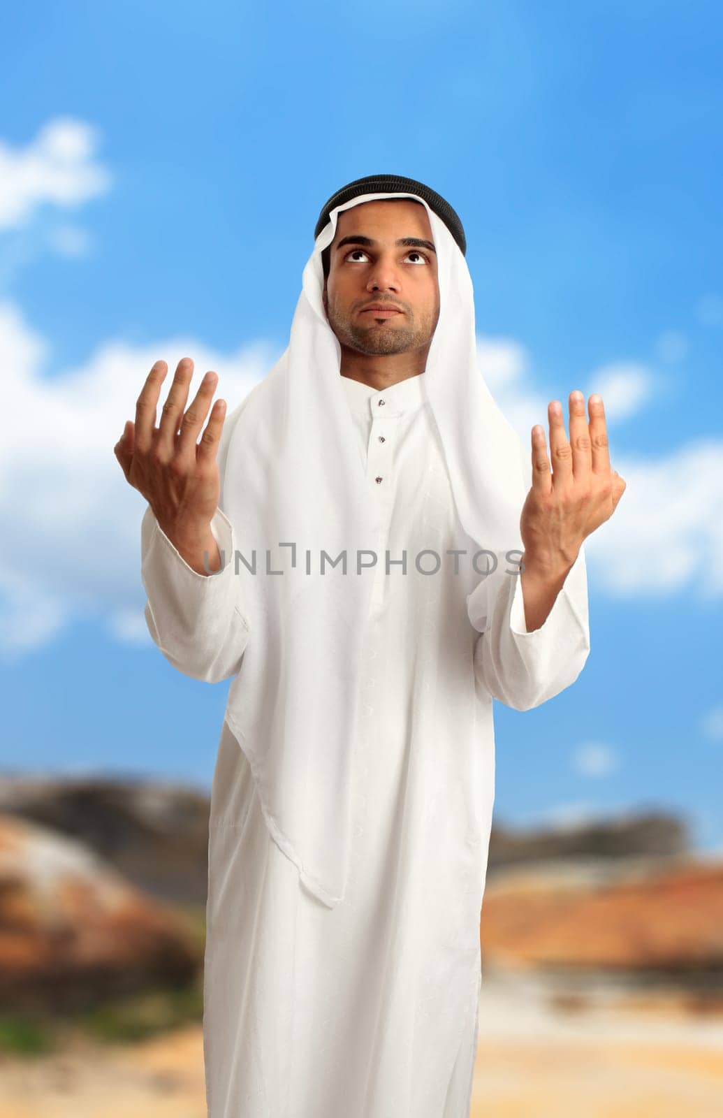 A mixed race man in an arid landscape, dressed in middle eastern clothing has arms outstretched in form of pleading, supplication, or prayer.