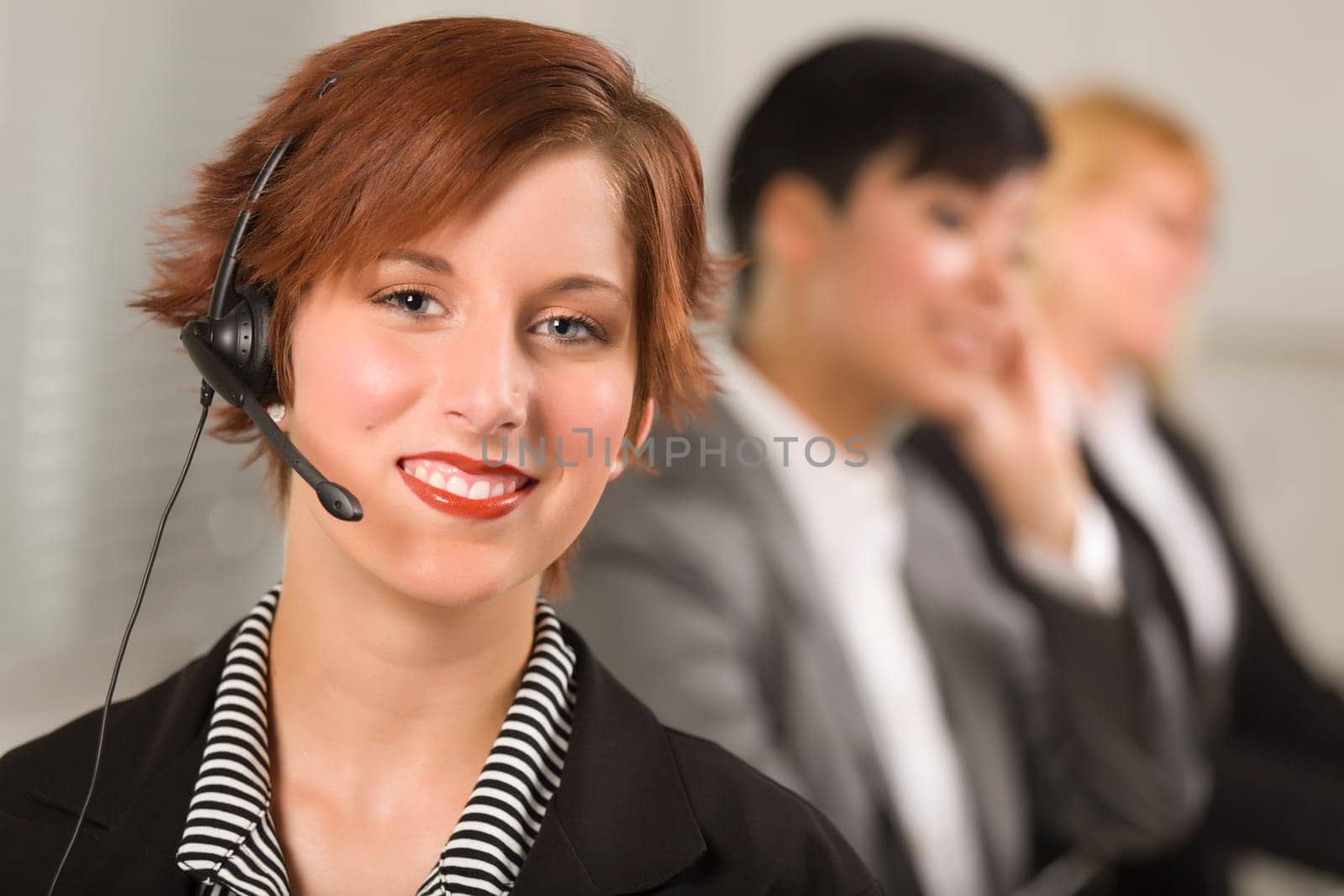 Pretty Red Haired Businesswoman with Headset and Colleagues Behind in Office Setting.