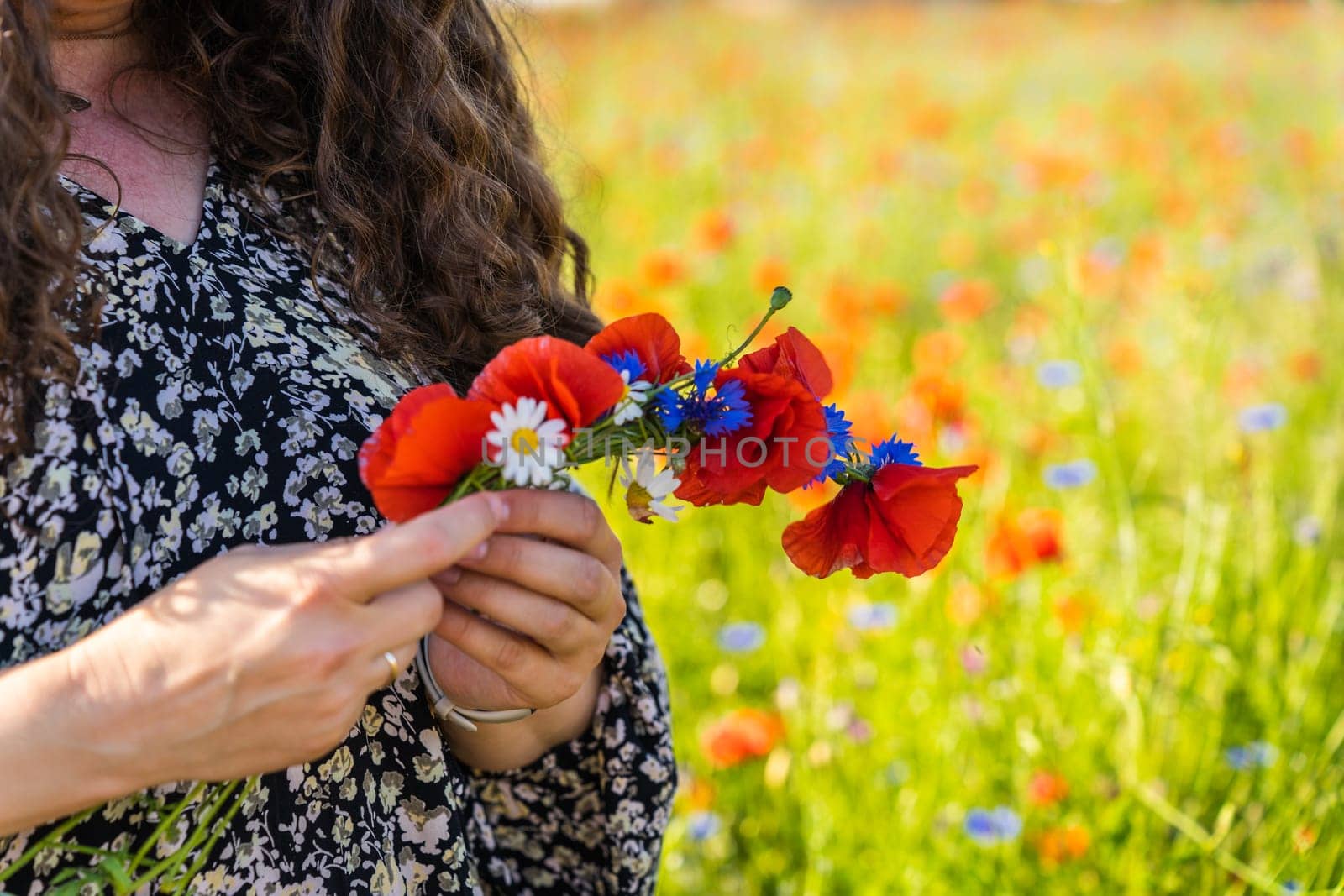 A long-haired curly woman weaves a wreath of wildflowers, poppies, daisies and cornflowers.