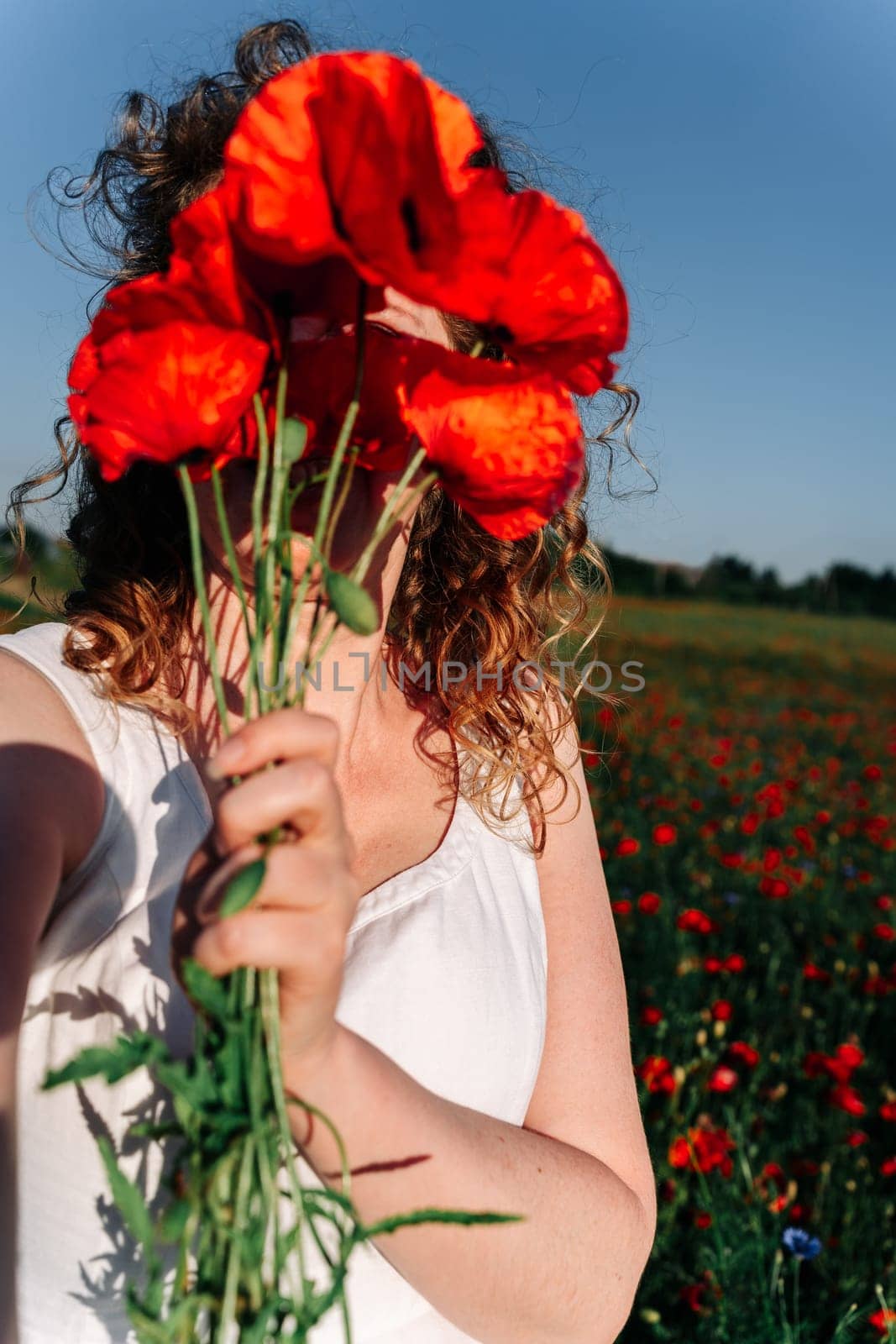 Selfie of a curly red-haired woman covering her face with a bunch of red field poppies among a poppy field.