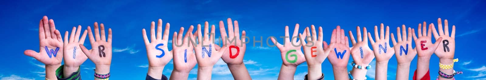 Children Hands Building Colorful German Word Wir Sind Gewinner Means We Are Winners. Blue Sky As Background